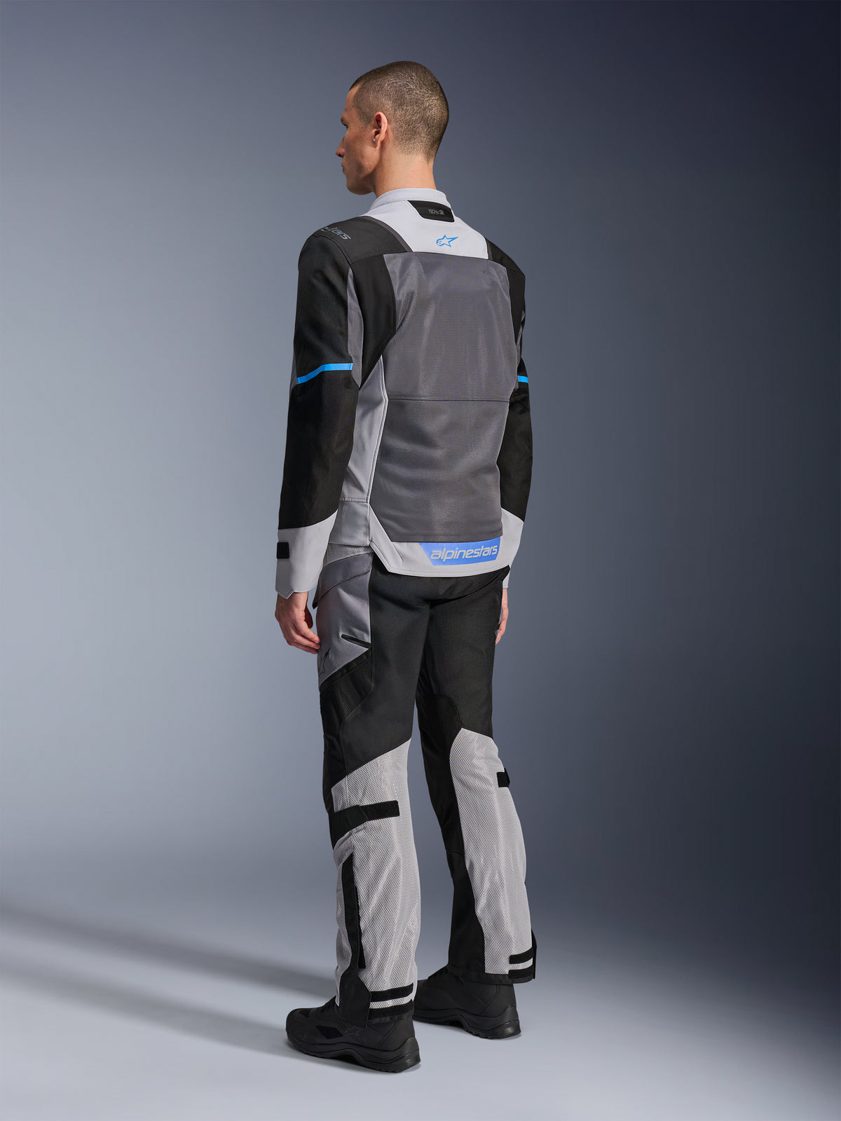 Veste Alpinestars ST-2 Air, modèle masculin vu de dos, coloris Ice Gray, Dark Gray et Black, dotée d'un grand panneau dorsal en mesh gris, accents de logo bleus et pantalon de moto assorti pour le touring estival.