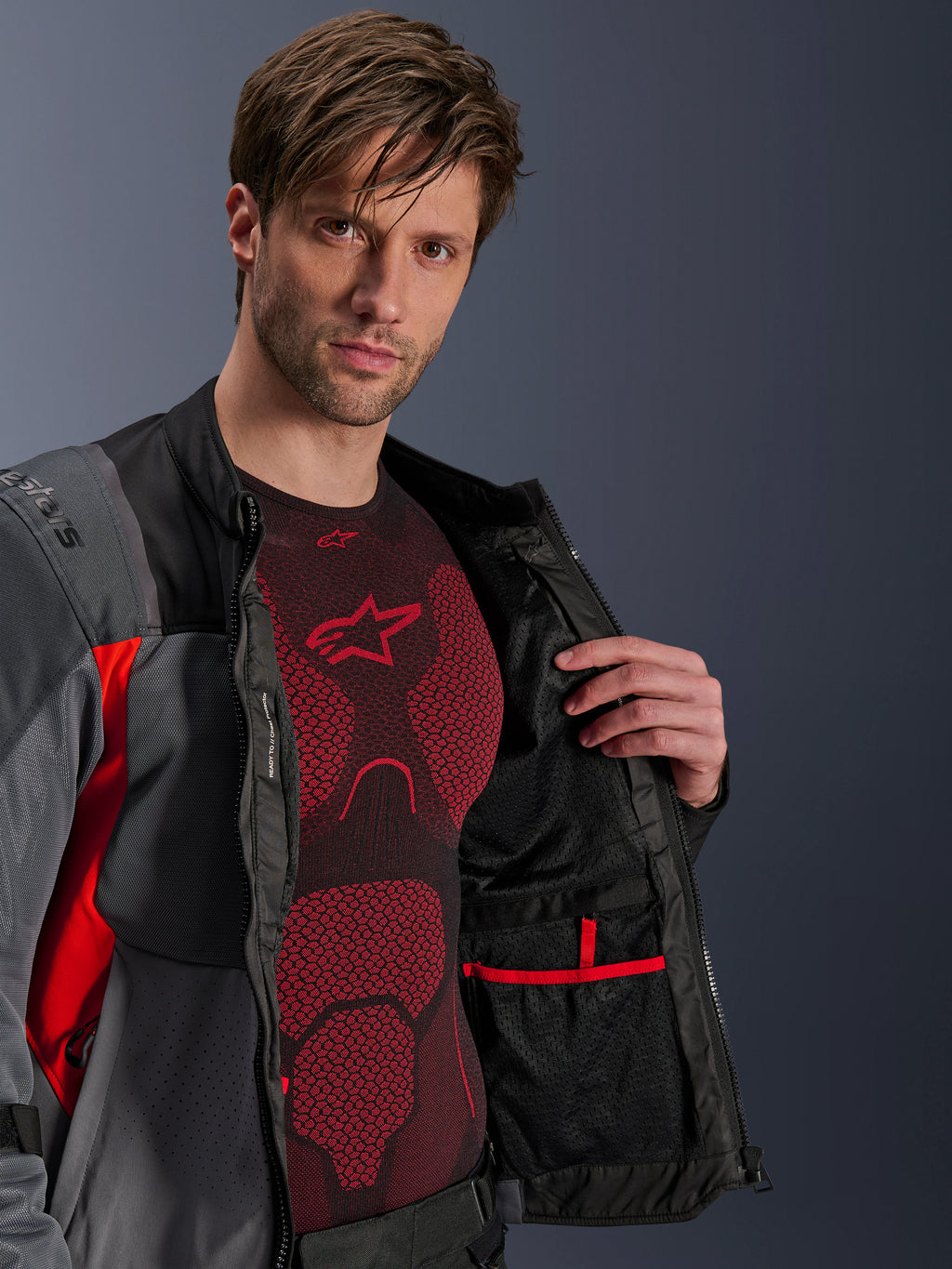 Veste Alpinestars ST-2 Air, gris foncé, gris anthracite et rouge vif, dézippée pour montrer la doublure intérieure en mesh et la poche bordée de rouge, portée sur une couche de base technique rouge, équipement de touring moto d'aventure moderne