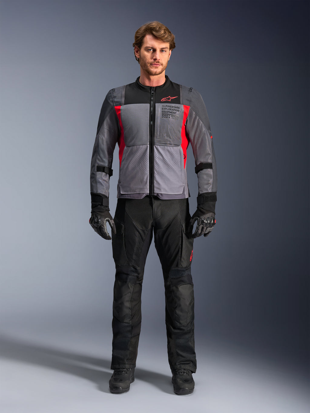 Veste Alpinestars ST-2 Air en gris foncé et rouge vif, portée par un mannequin homme avec un pantalon et des gants de moto noirs, dotée de larges panneaux en maille pour la circulation de l'air et d'un marquage moderne d'équipement d'exploration