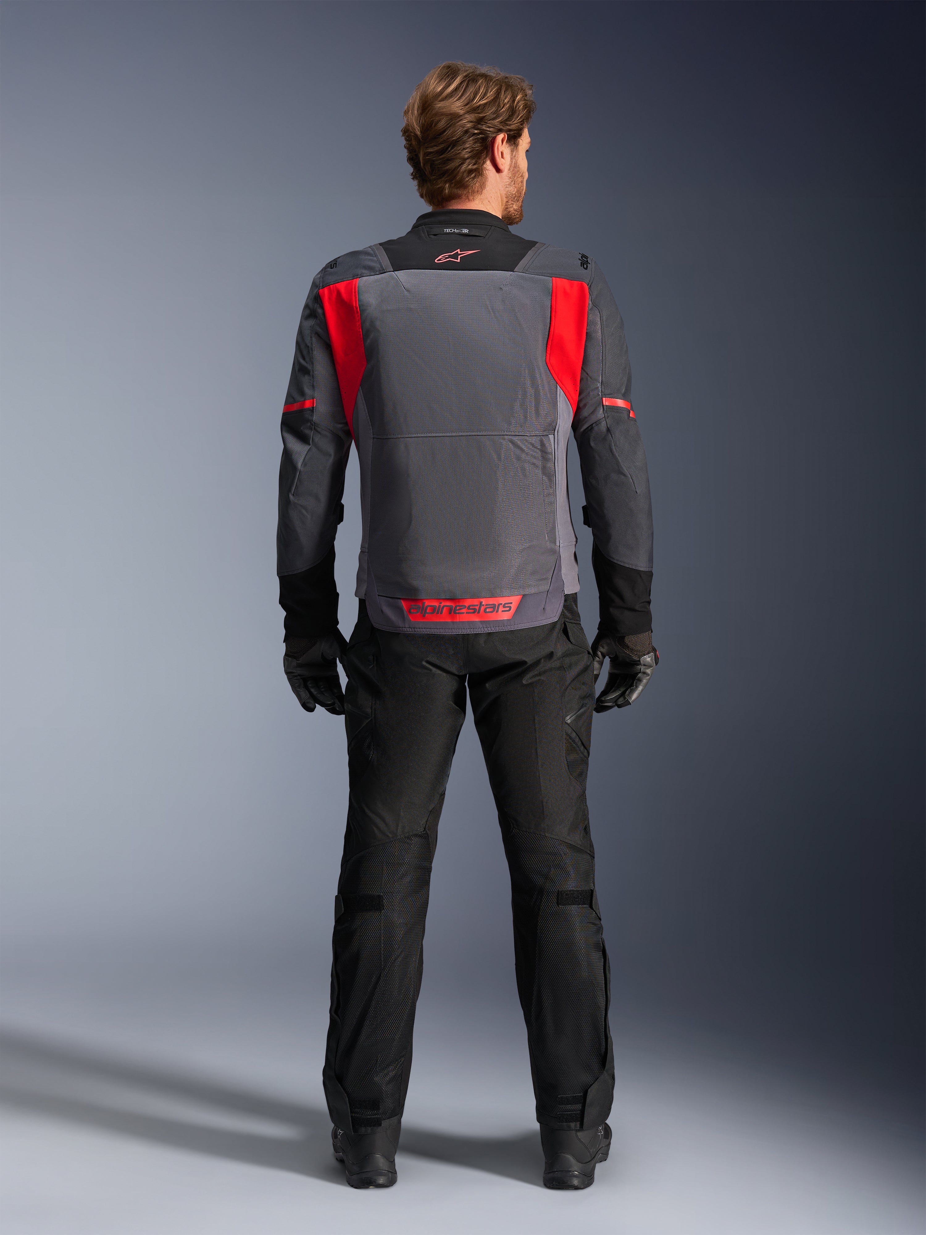 Veste Alpinestars ST-2 Air, Gris Profond, Gris Foncé et Rouge Éclatant, vue de dos portée par un motard avec pantalon et gants noirs, dotée de grands panneaux de ventilation en mesh et d'un logo Alpinestars rouge dans le bas du dos, design de touring estival moderne