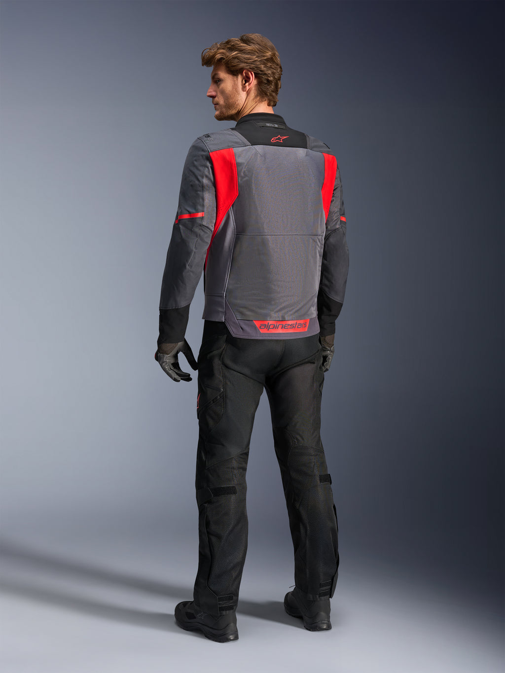 Motard vu de dos portant la veste Alpinestars St-2 Air en gris foncé, gris anthracite et rouge vif, mettant en valeur le grand panneau de ventilation en mesh à l'arrière et les marquages rouges, assortie d'un pantalon de moto noir et de gants gris.
