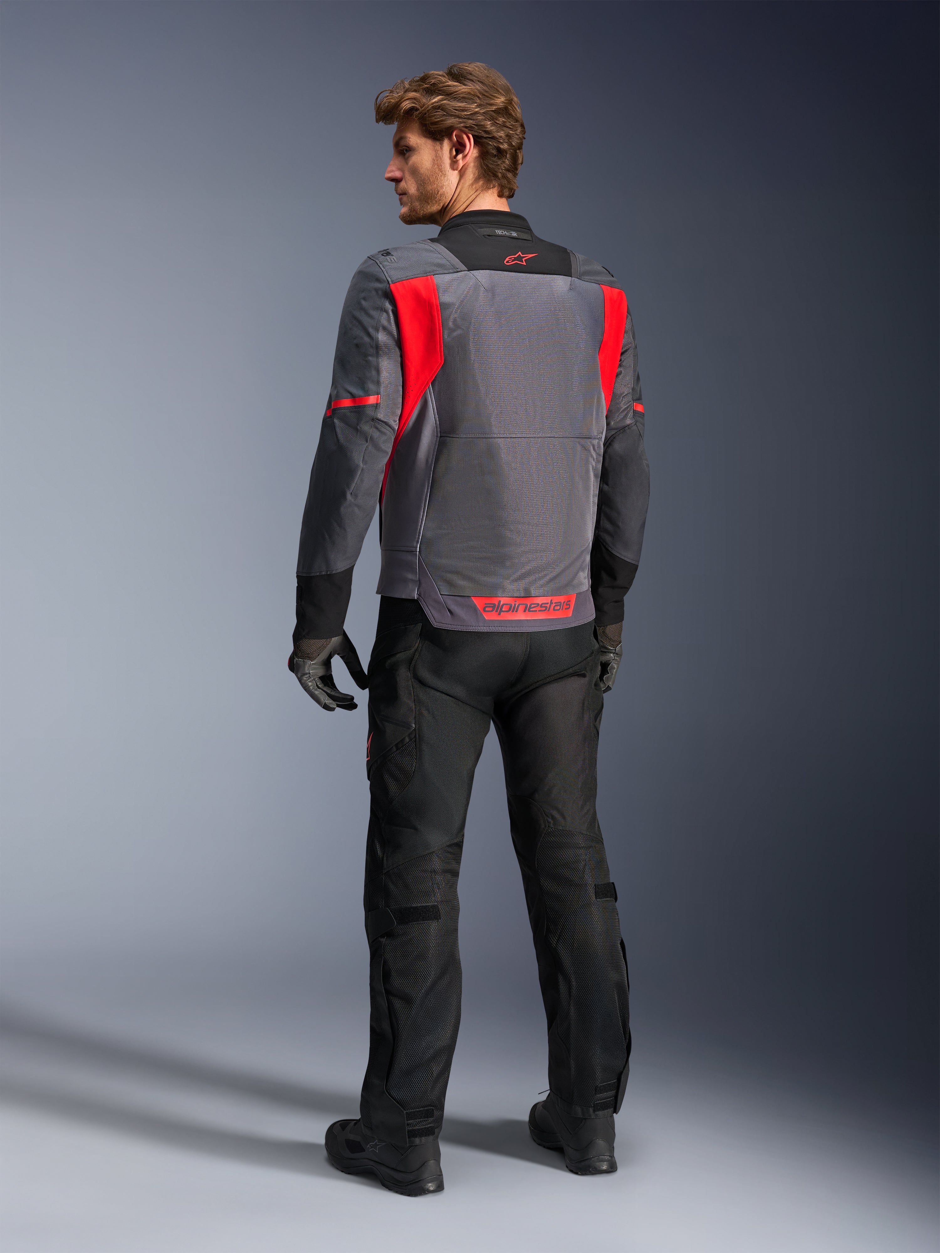 Motard vu de dos portant la veste Alpinestars St-2 Air en gris foncé, gris anthracite et rouge vif, mettant en valeur le grand panneau de ventilation en mesh à l'arrière et les marquages rouges, assortie d'un pantalon de moto noir et de gants gris.