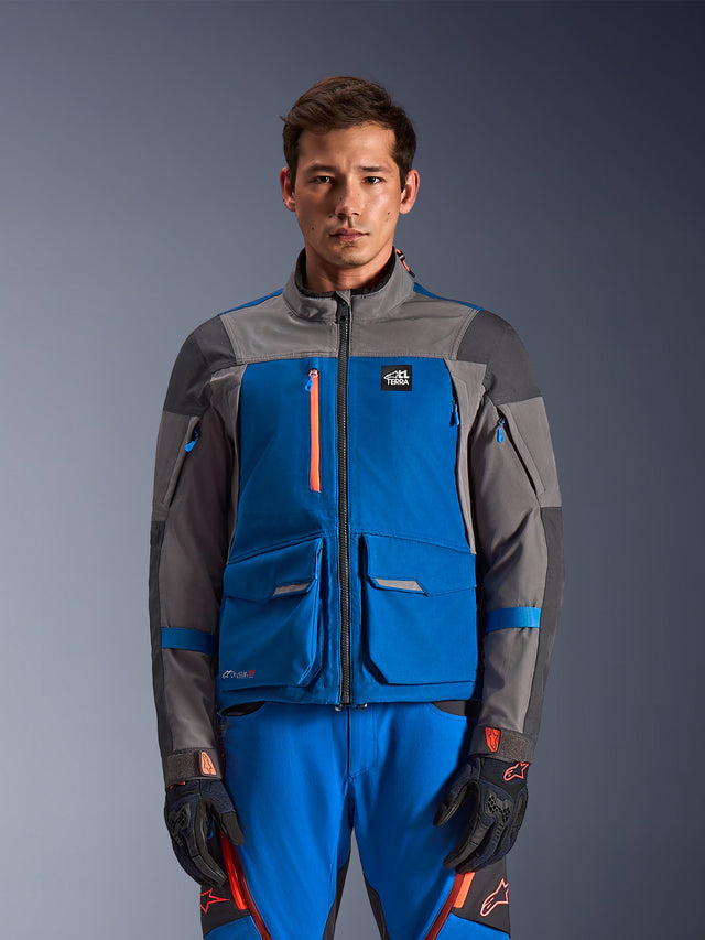 Alpinestars Maxdura Drystar®XF Laminated Armored Shell, Adventure Motorcycle Jacket, Royal Blue and Dark Gray, worn with matching blue pants and black gloves with orange accents, features multi-pocket design, orange chest zipper, and 'All Terra' branding, rugged technical look for adventure riding