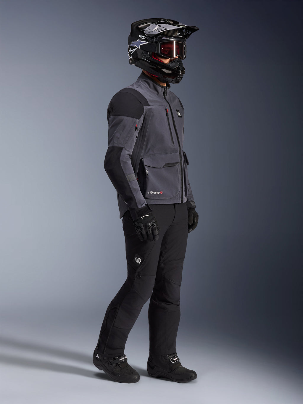 Motorcycle rider wearing Alpinestars Maxdura Drystar®XF Laminated Armored Shell jacket in Deep Gray and Black, paired with black adventure riding pants, black helmet with goggles, black gloves and boots, full-body studio shot highlighting the technical dual-sport gear.