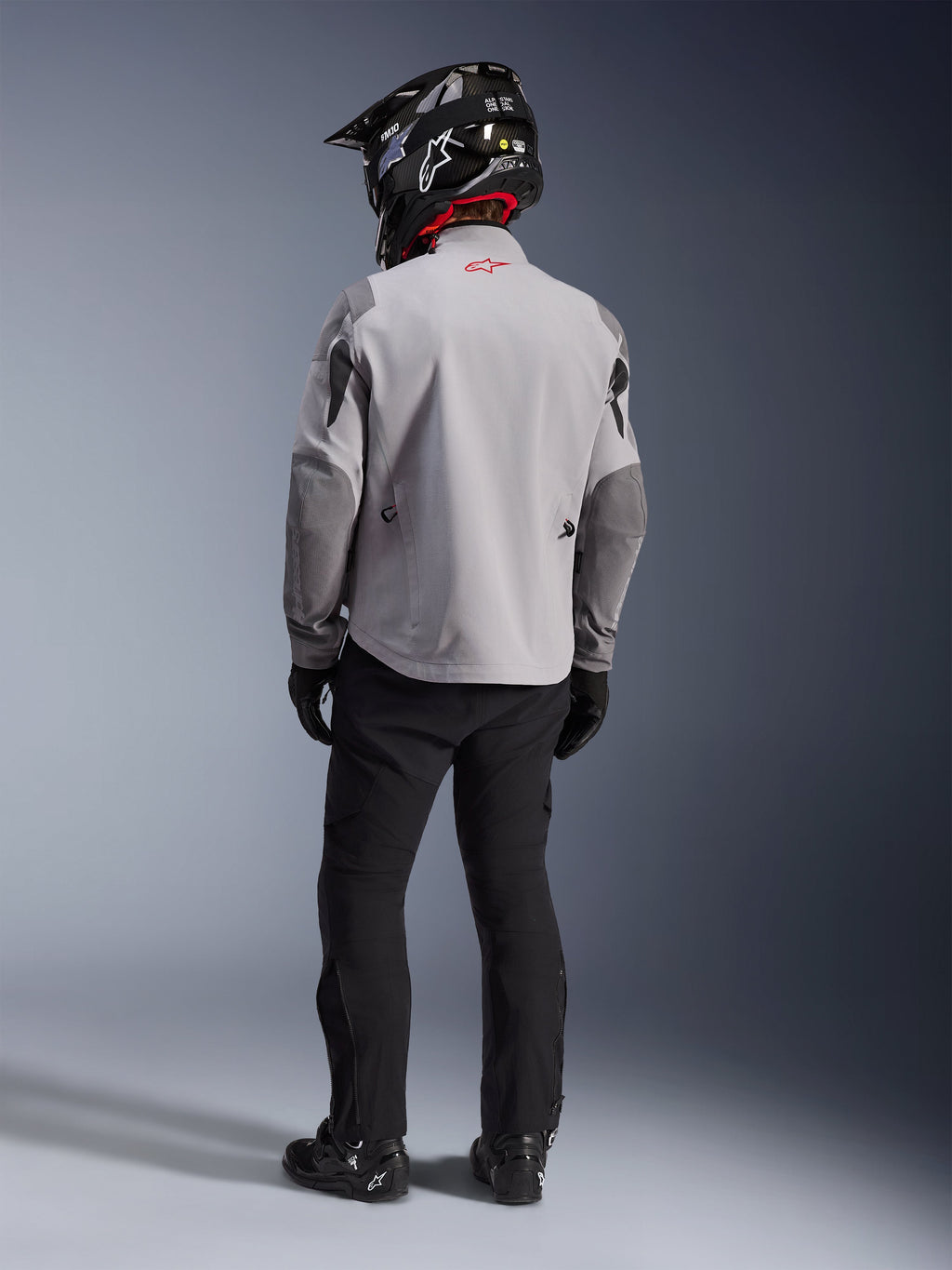 Rear view of a motorcycle rider wearing the Alpinestars Maxdura Drystar®XF Laminated Armored Shell jacket in Mid Gray and Dark Gray, featuring a red star logo on the collar, paired with black adventure pants, a black carbon-patterned helmet, and black boots.