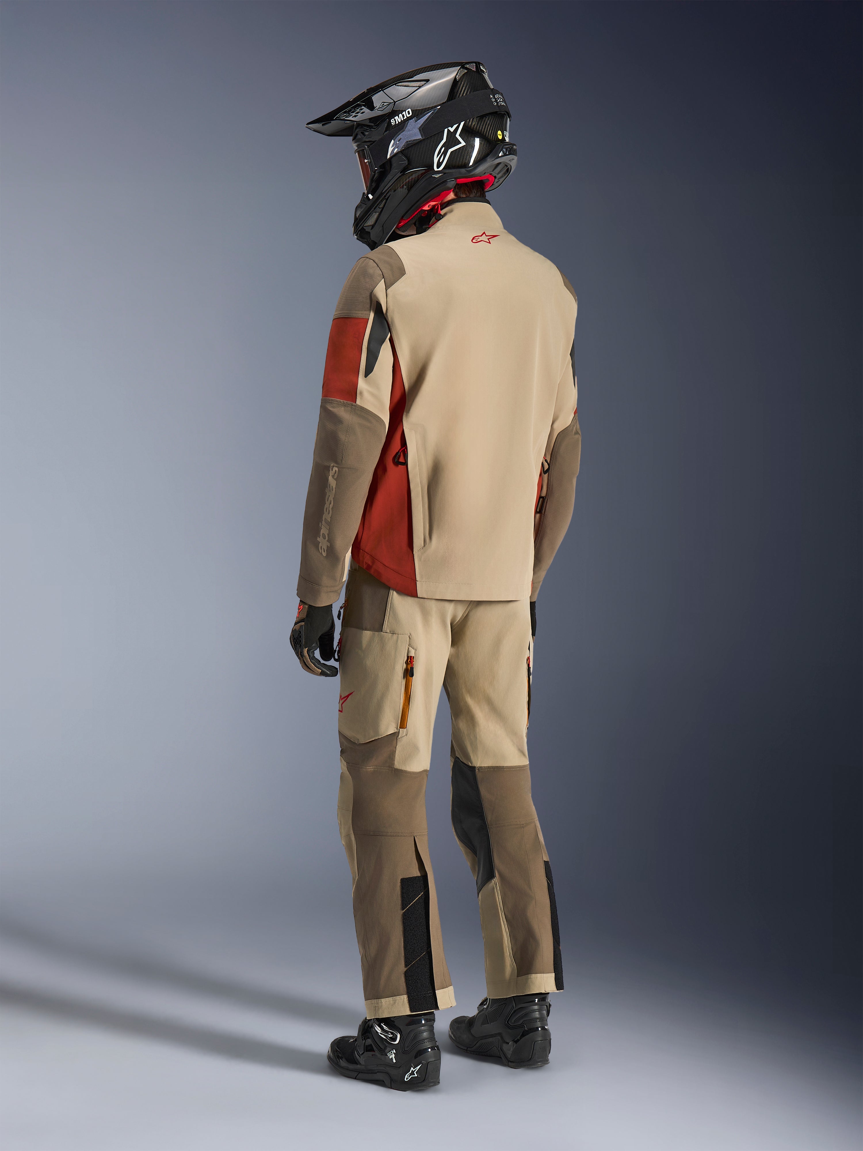 Adventure rider from the back wearing the Alpinestars Maxdura Drystar®XF Laminated Armored Shell in Aluminum Walnut Rust Brown, paired with matching riding pants, black off-road helmet, and black boots, complete technical touring outfit.