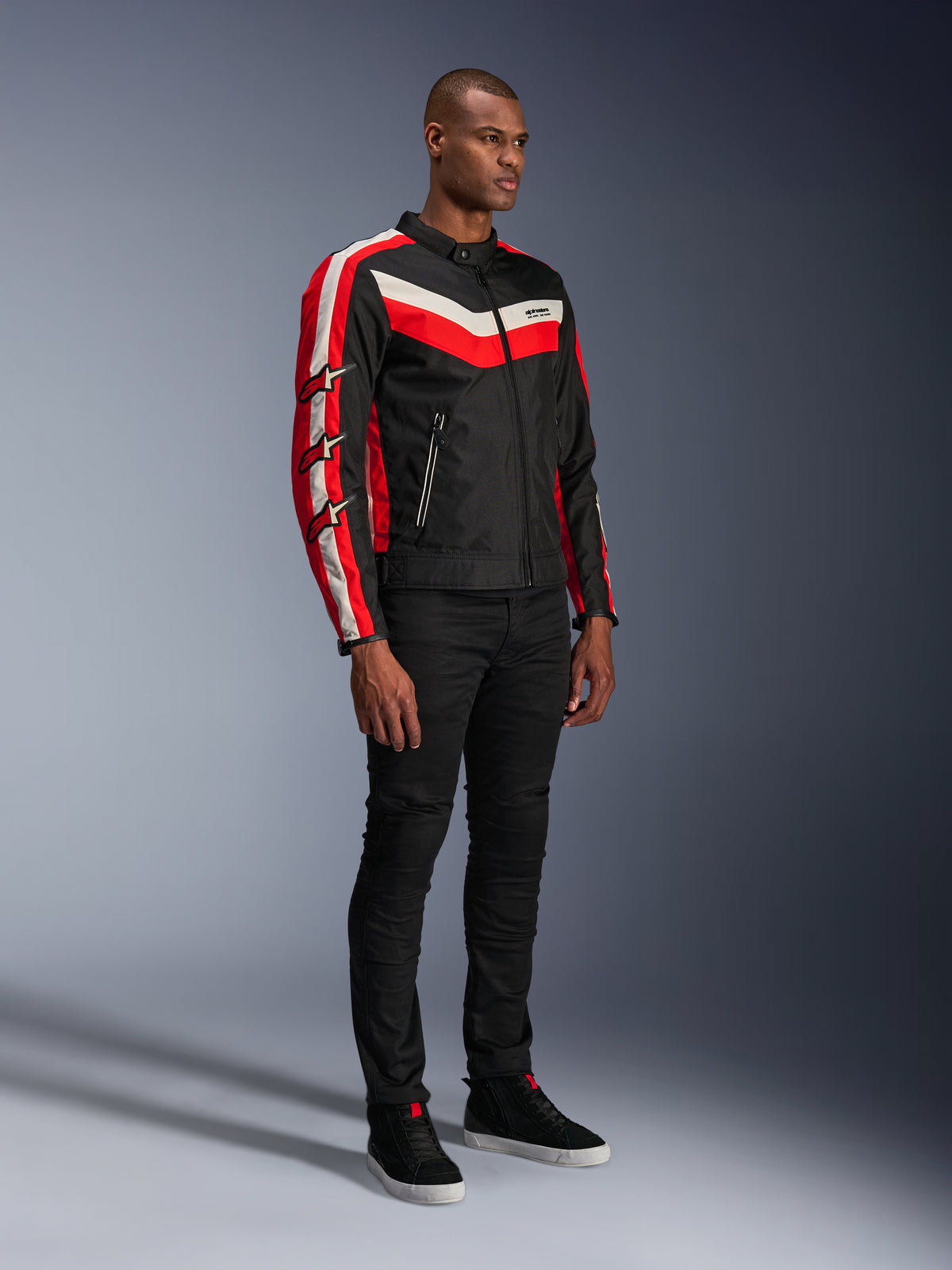 Modèle masculin portant la veste Alpinestars T-Dyno WR, veste de moto en textile noir, rouge éclatant et écru, avec rayures rouges et blanc cassé et logos étoiles Alpinestars sur les manches, accompagnée d'un pantalon noir et de baskets montantes noires, équipement de moto urbain d'inspiration vintage.