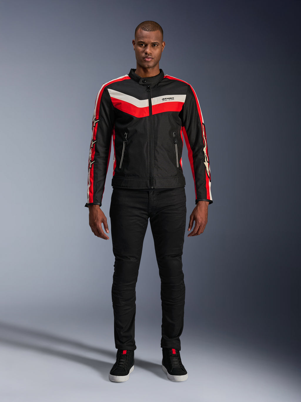 Veste Alpinestars T-Dyno WR, veste de moto en textile noir, rouge vif et écru, avec des bandes en V sur la poitrine et des logos étoilés sur les manches, portée avec un pantalon noir et des chaussures de moto noires, équipement urbain d'inspiration héritage