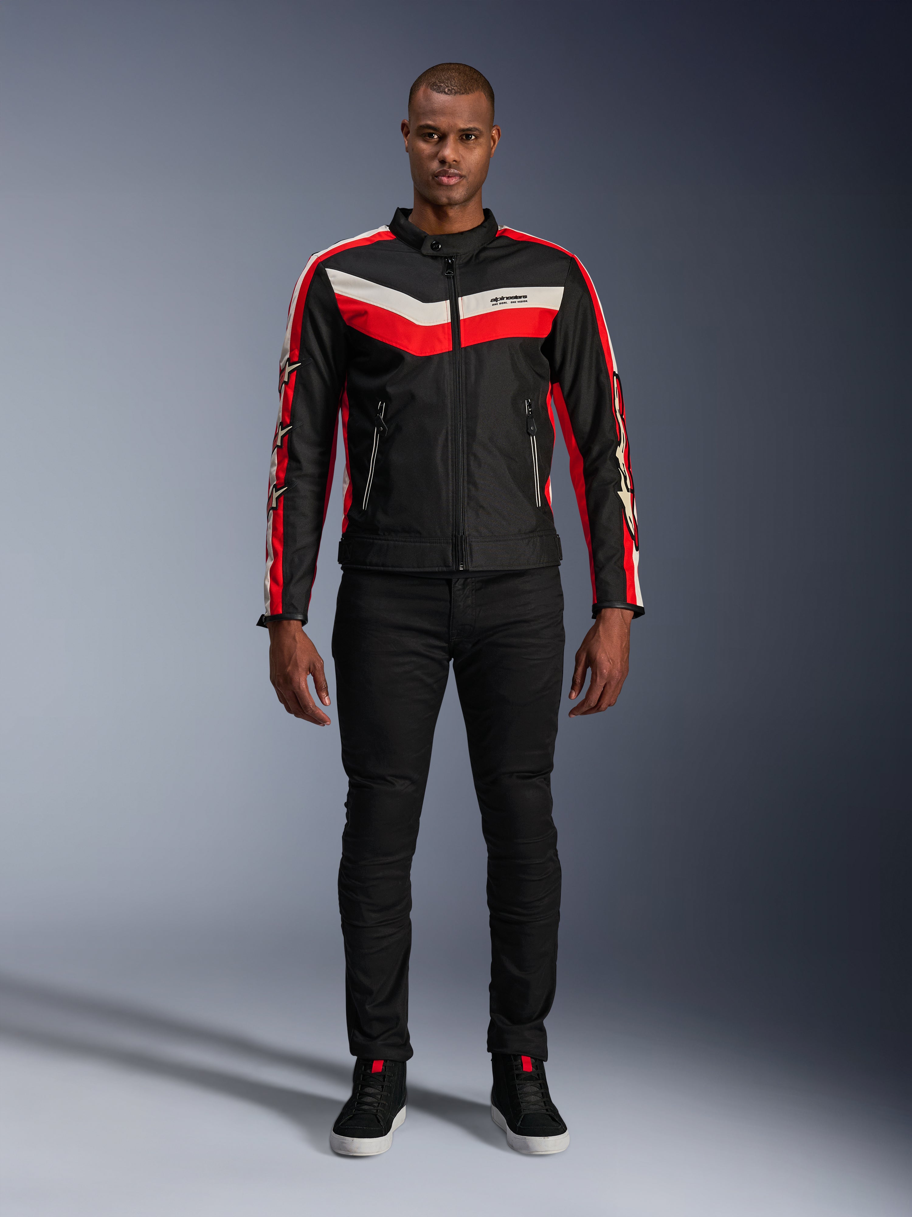 Veste Alpinestars T-Dyno WR, veste de moto en textile noir, rouge vif et écru, avec des bandes en V sur la poitrine et des logos étoilés sur les manches, portée avec un pantalon noir et des chaussures de moto noires, équipement urbain d'inspiration héritage