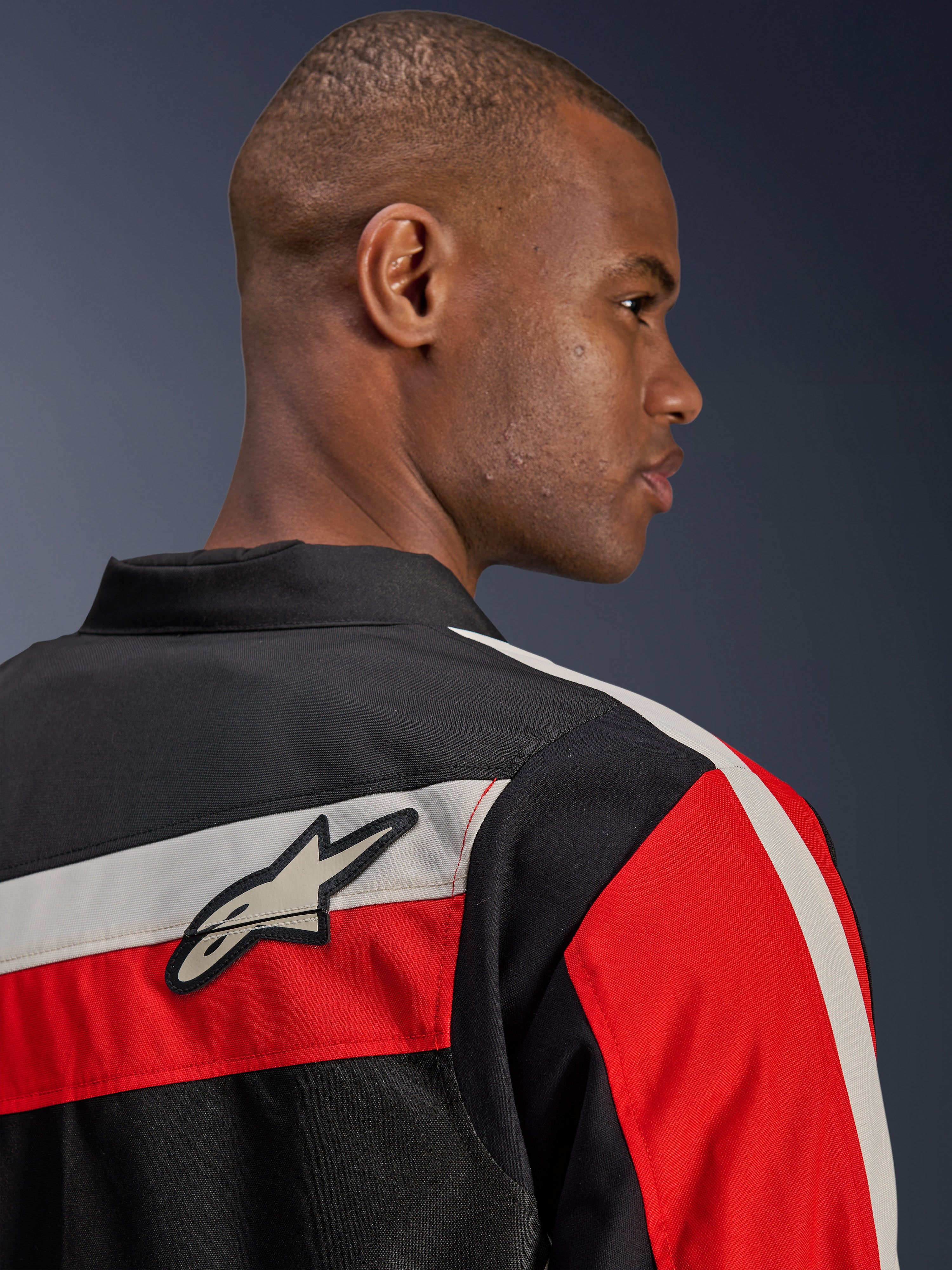 Veste Alpinestars T-Dyno WR, vue arrière en gros plan sur un mannequin, veste de moto en textile noir avec bandes horizontales écrues et rouge vif, ornée du patch logo étoilé Alpinestars sur l'épaule, style biker d'inspiration héritage