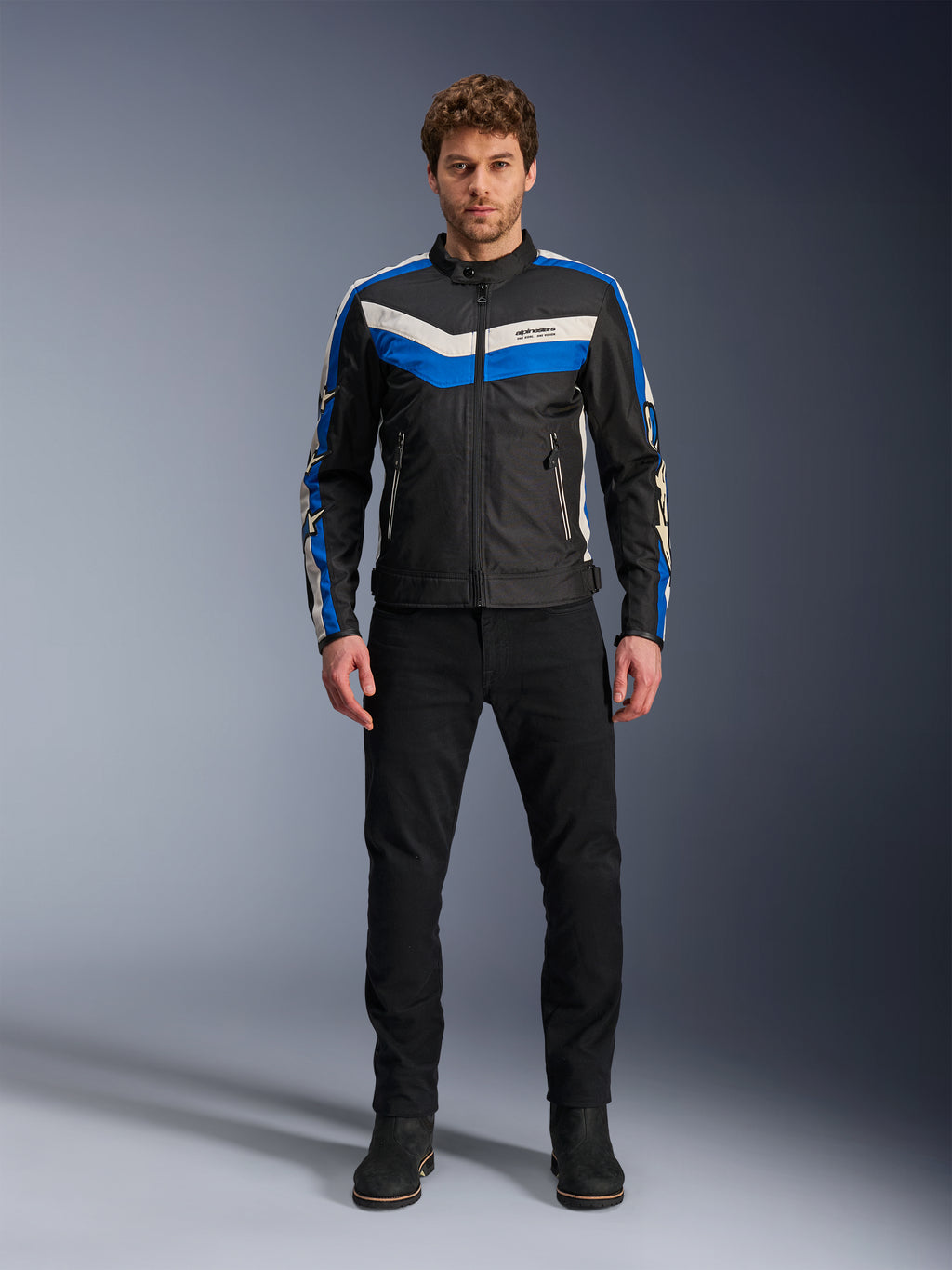Veste Alpinestars T-Dyno WR, veste de moto en textile, noir, bleu et écru, portée par un mannequin homme avec un pantalon et des bottes noirs, arborant des bandes en chevron sur la poitrine et un design biker héritage