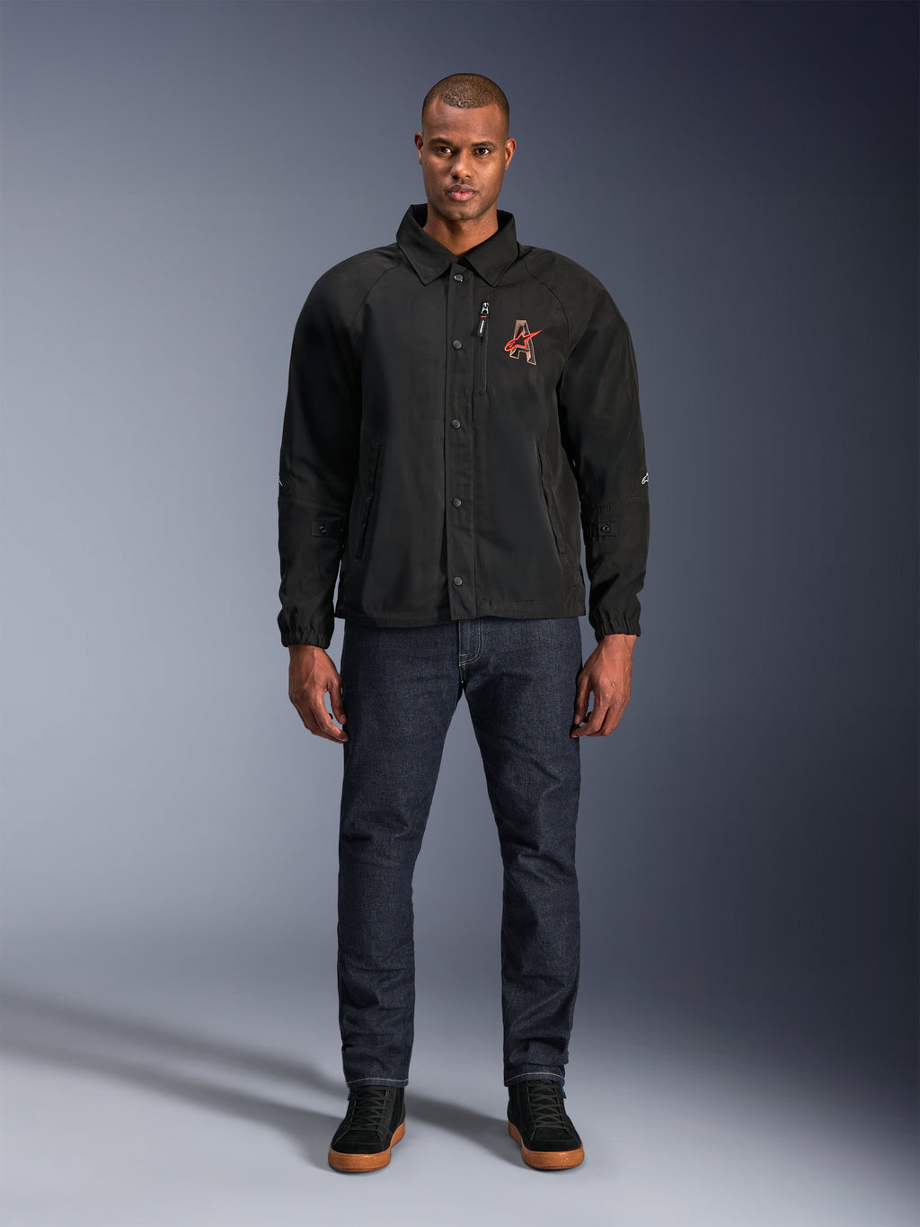 Veste Alpinestars Revved WR noire, portée par un mannequin avec un jean bleu foncé et des baskets noires, vue de face, dotée d'une fermeture par boutons-pression et d'un logo brodé en 3D sur la poitrine, équipement de moto urbain contemporain.