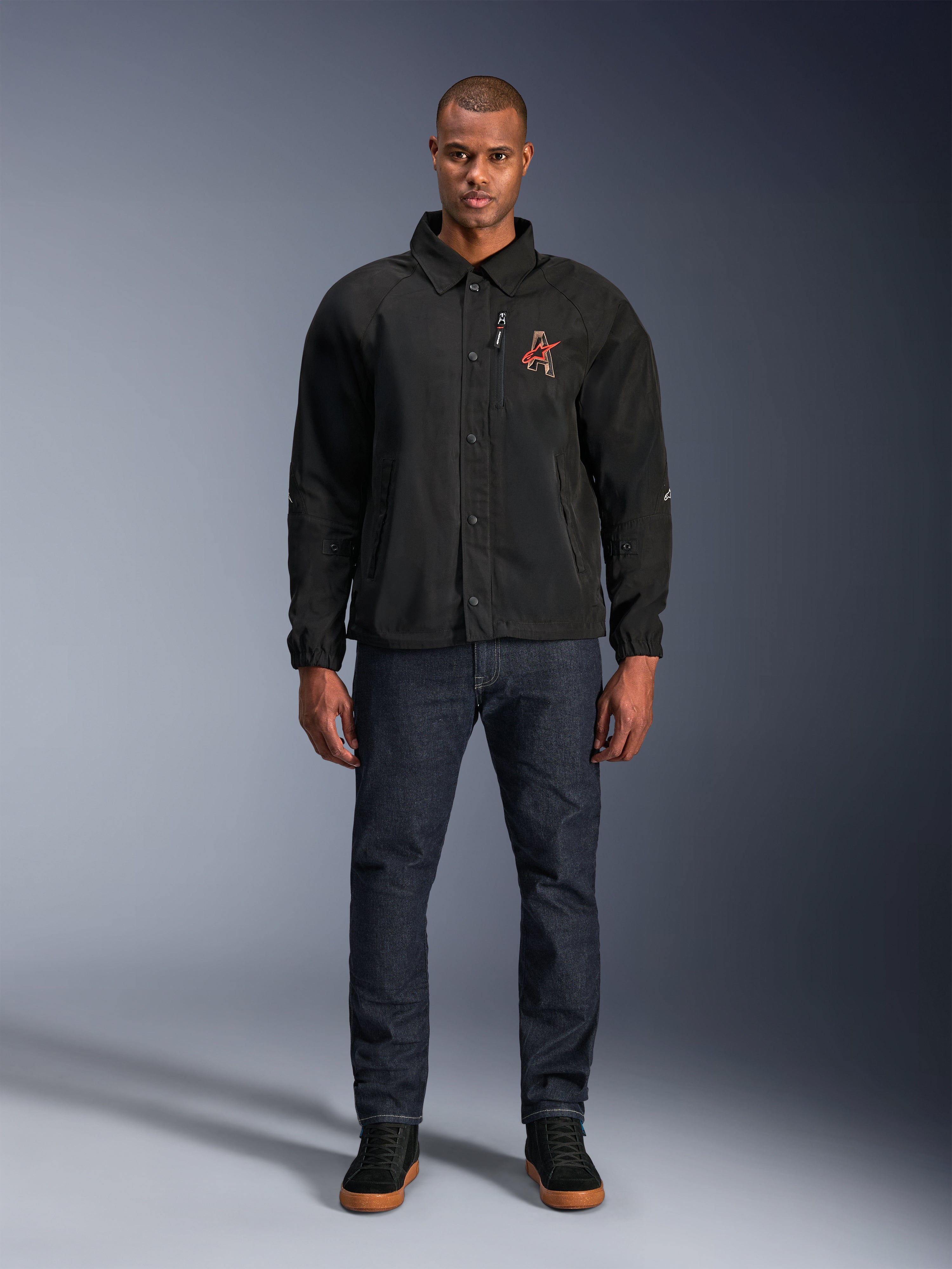 Veste Alpinestars Revved WR noire, portée par un mannequin avec un jean bleu foncé et des baskets noires, vue de face, dotée d'une fermeture par boutons-pression et d'un logo brodé en 3D sur la poitrine, équipement de moto urbain contemporain.
