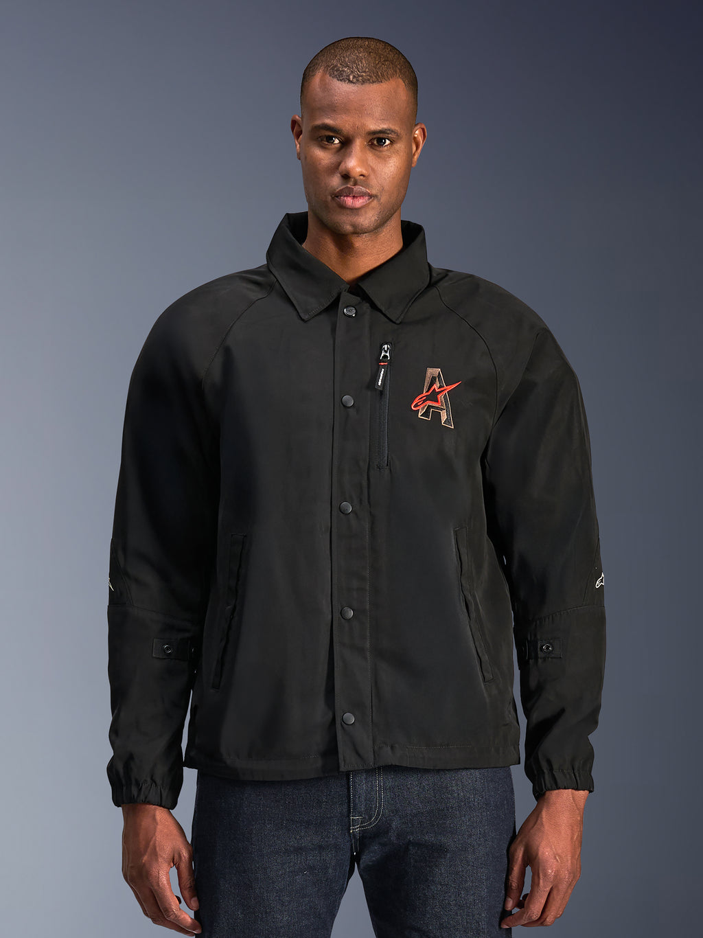 Veste Alpinestars Revved WR, veste moto noire, avec col style chemise et boutons-pression sur le devant, logo 'A' rouge brodé sur la poitrine, poche de poitrine zippée verticale, poignets élastiques, design de veste de coach urbaine