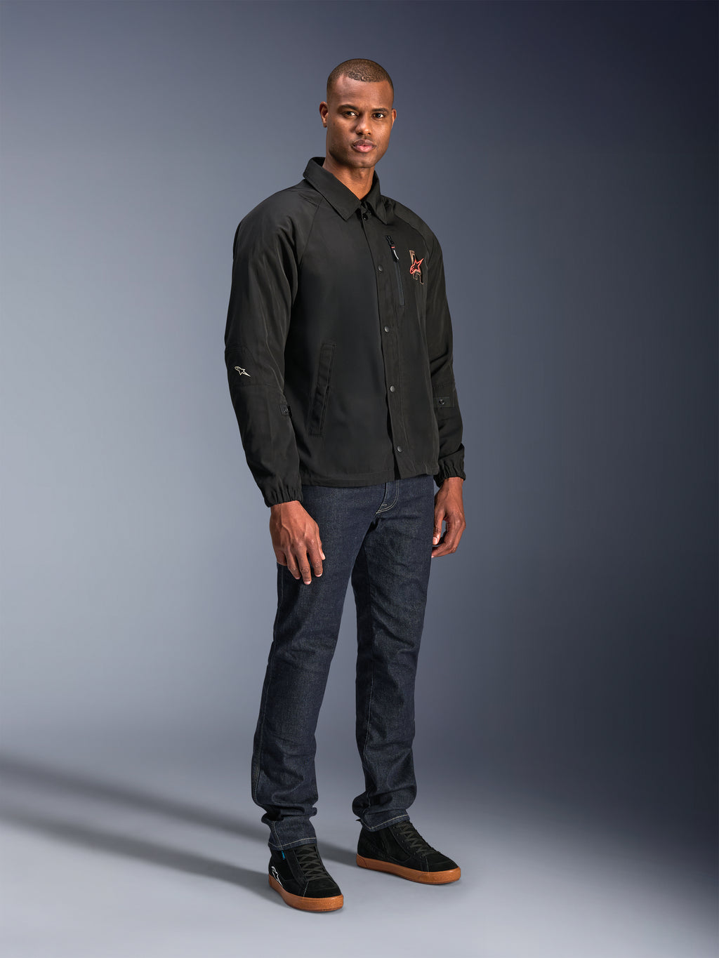 Veste Alpinestars Revved WR en noir, veste de moto urbaine style coach portée par un mannequin avec un jean bleu foncé et des baskets noires, dotée d'une fermeture à boutons-pression et d'un logo brodé, design minimaliste et sportif pour la conduite sur route.