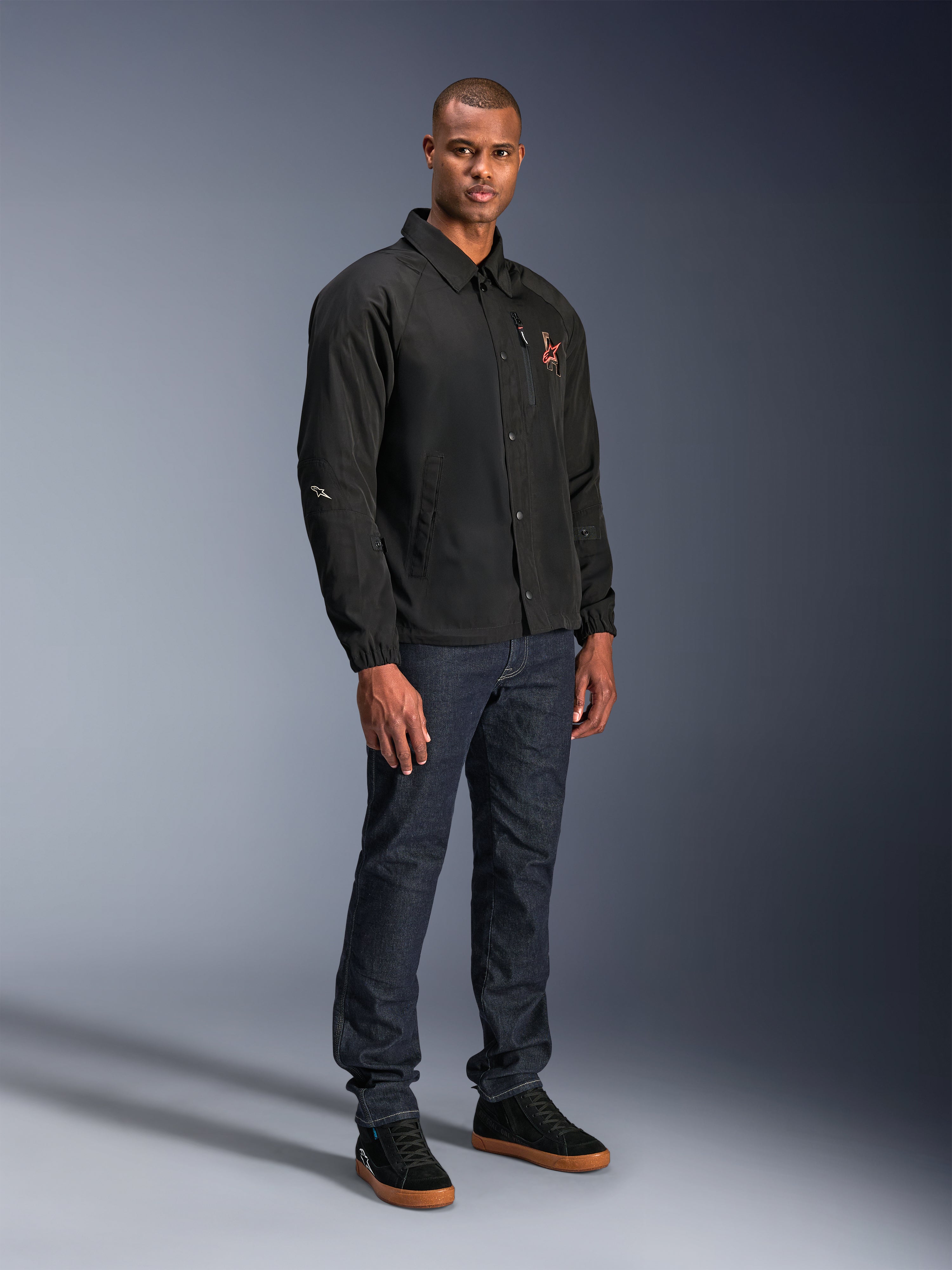 Veste Alpinestars Revved WR en noir, veste de moto urbaine style coach portée par un mannequin avec un jean bleu foncé et des baskets noires, dotée d'une fermeture à boutons-pression et d'un logo brodé, design minimaliste et sportif pour la conduite sur route.