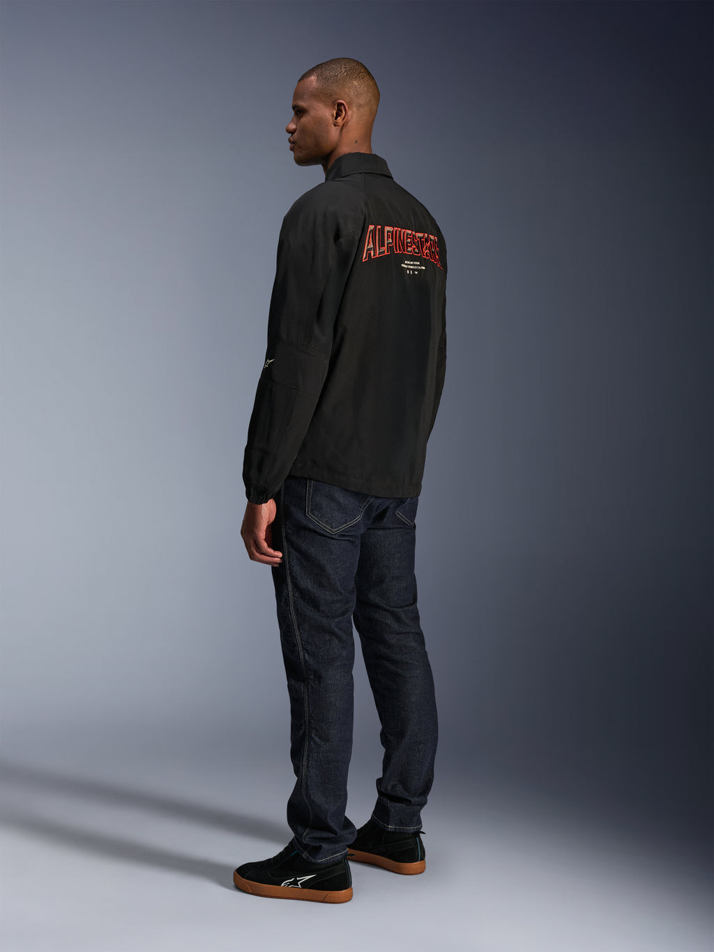 Veste Alpinestars Revved WR, noire, avec un grand logo Alpinestars orange brodé en 3D au dos, vue arrière de trois-quarts d'un mannequin portant un jean bleu foncé et des baskets noires, veste de moto urbaine contemporaine avec une silhouette de veste de coach