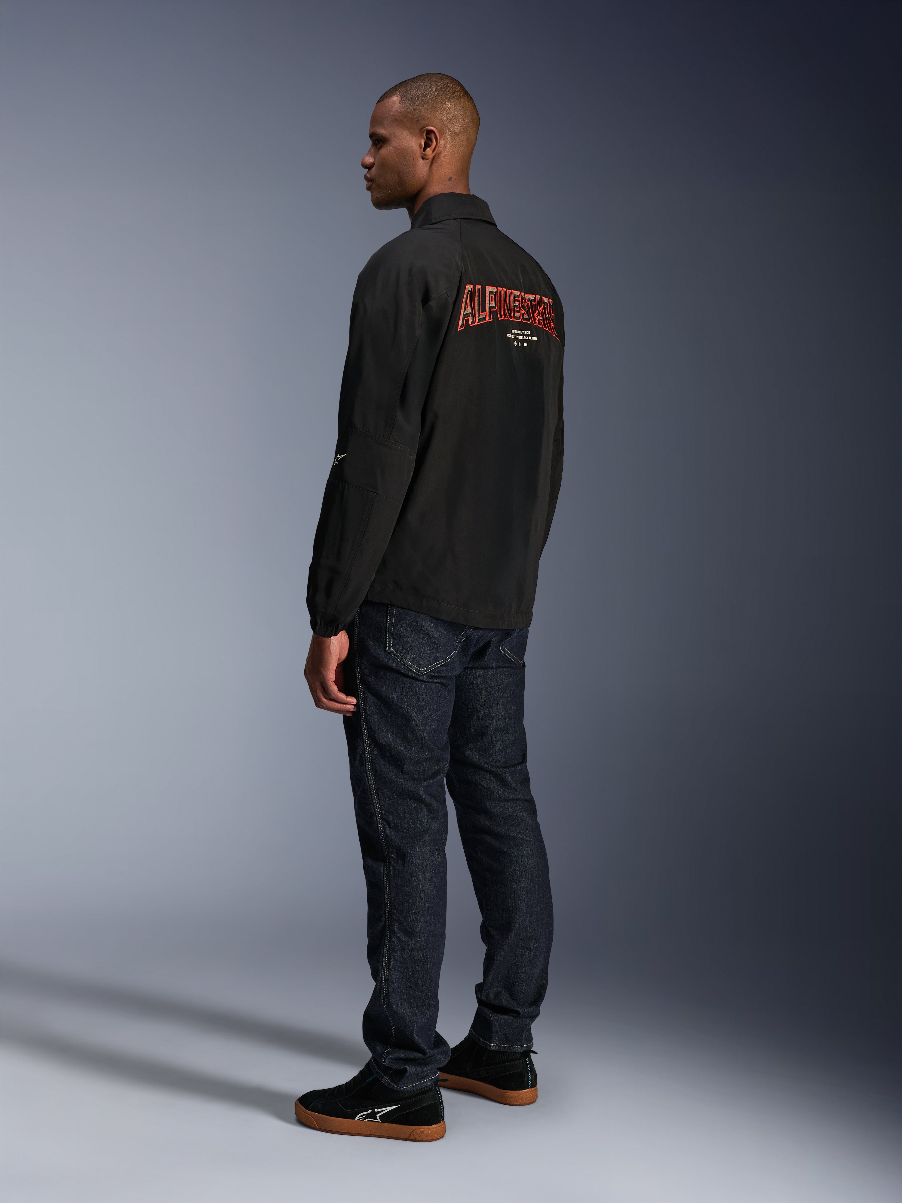 Veste Alpinestars Revved WR, noire, avec un grand logo Alpinestars orange brodé en 3D au dos, vue arrière de trois-quarts d'un mannequin portant un jean bleu foncé et des baskets noires, veste de moto urbaine contemporaine avec une silhouette de veste de coach