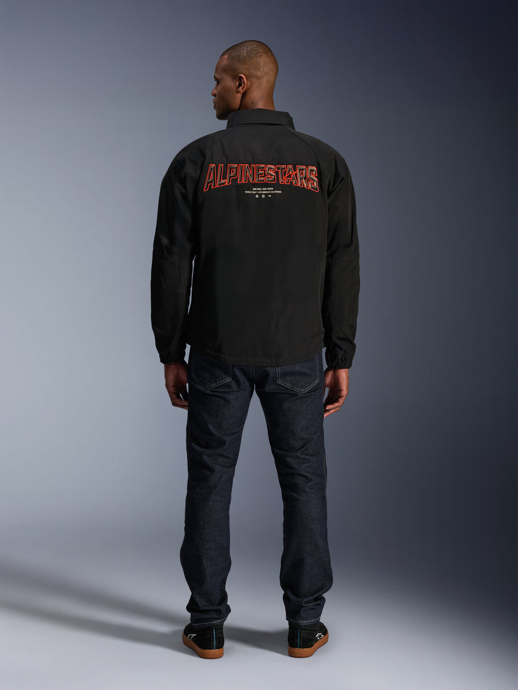 Veste Alpinestars Revved WR, noire, vue de dos avec une grande broderie rouge « ALPINESTARS » à effet 3D, veste de style coach urbain, portée avec un jean en denim foncé et des baskets noires, vêtement de moto minimaliste