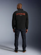 Veste Alpinestars Revved WR, noire, vue de dos avec une grande broderie rouge « ALPINESTARS » à effet 3D, veste de style coach urbain, portée avec un jean en denim foncé et des baskets noires, vêtement de moto minimaliste