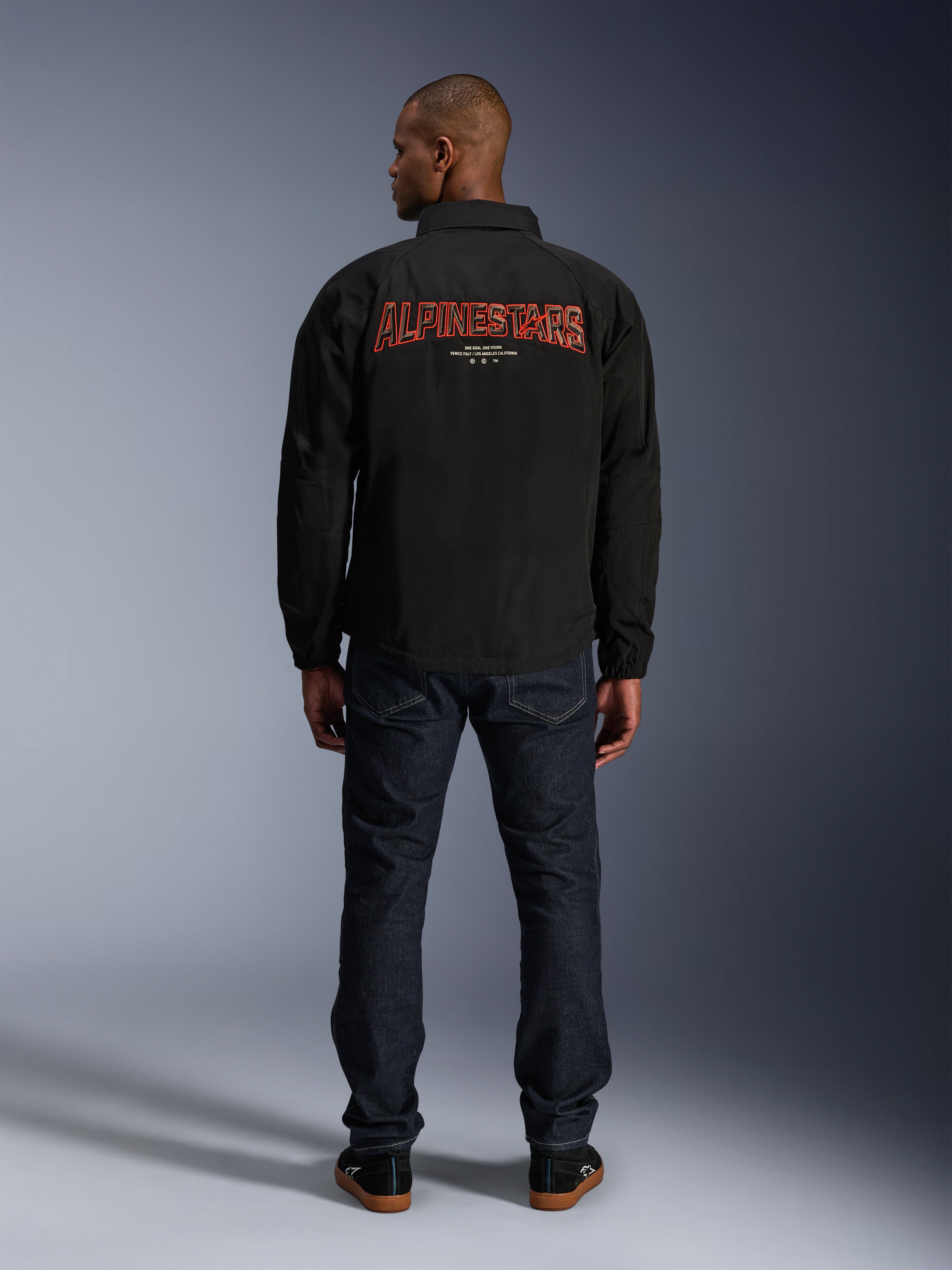 Veste Alpinestars Revved WR, noire, vue de dos avec une grande broderie rouge « ALPINESTARS » à effet 3D, veste de style coach urbain, portée avec un jean en denim foncé et des baskets noires, vêtement de moto minimaliste