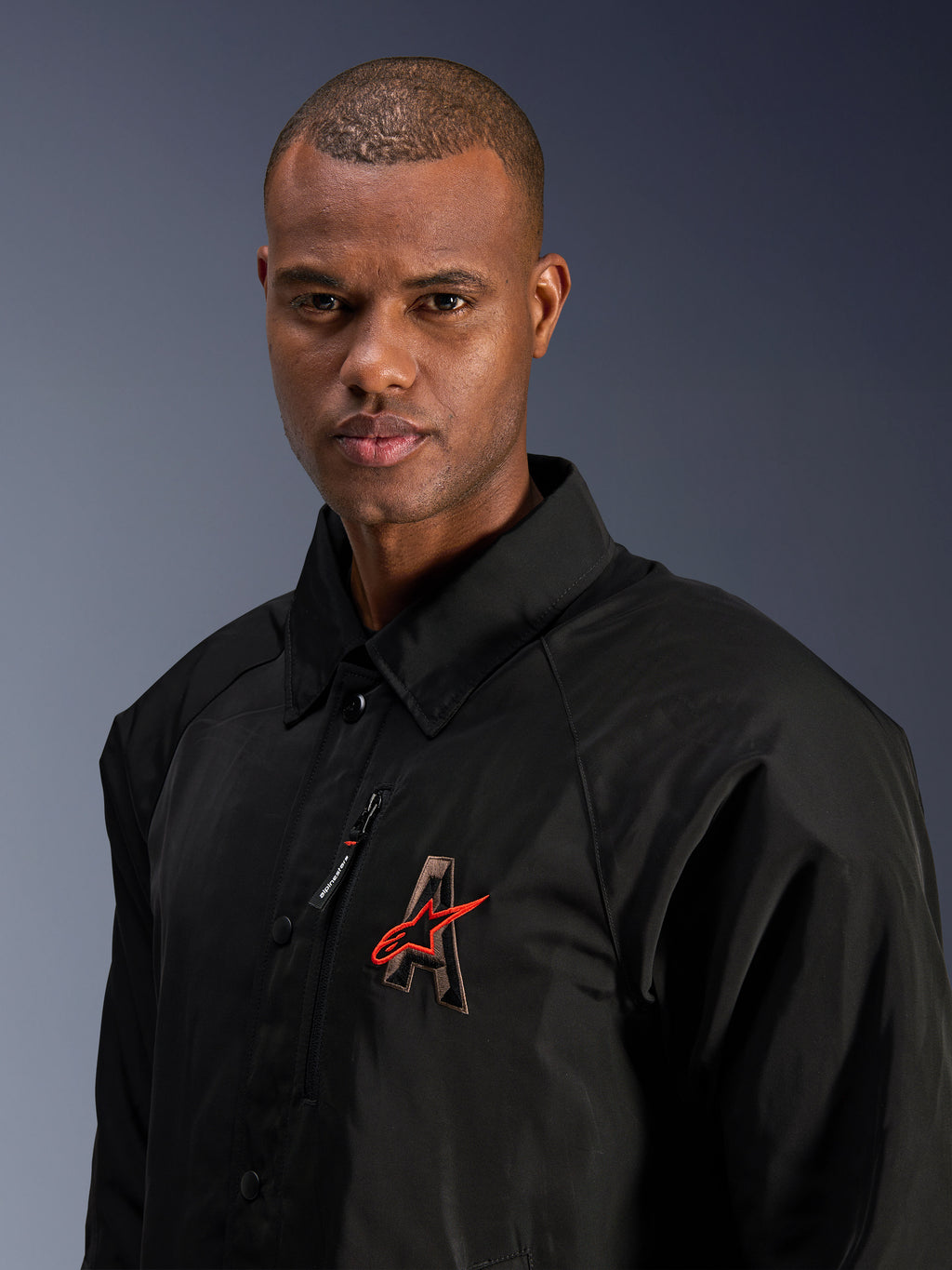 Veste Alpinestars Revved WR, noire, gros plan d'un homme portant la veste de moto en satin résistante à l'eau, avec le logo « A » brodé en 3D et l'étoile Alpinestars rouge, une poche de poitrine zippée et un design avec col pour le pilotage urbain contemporain.