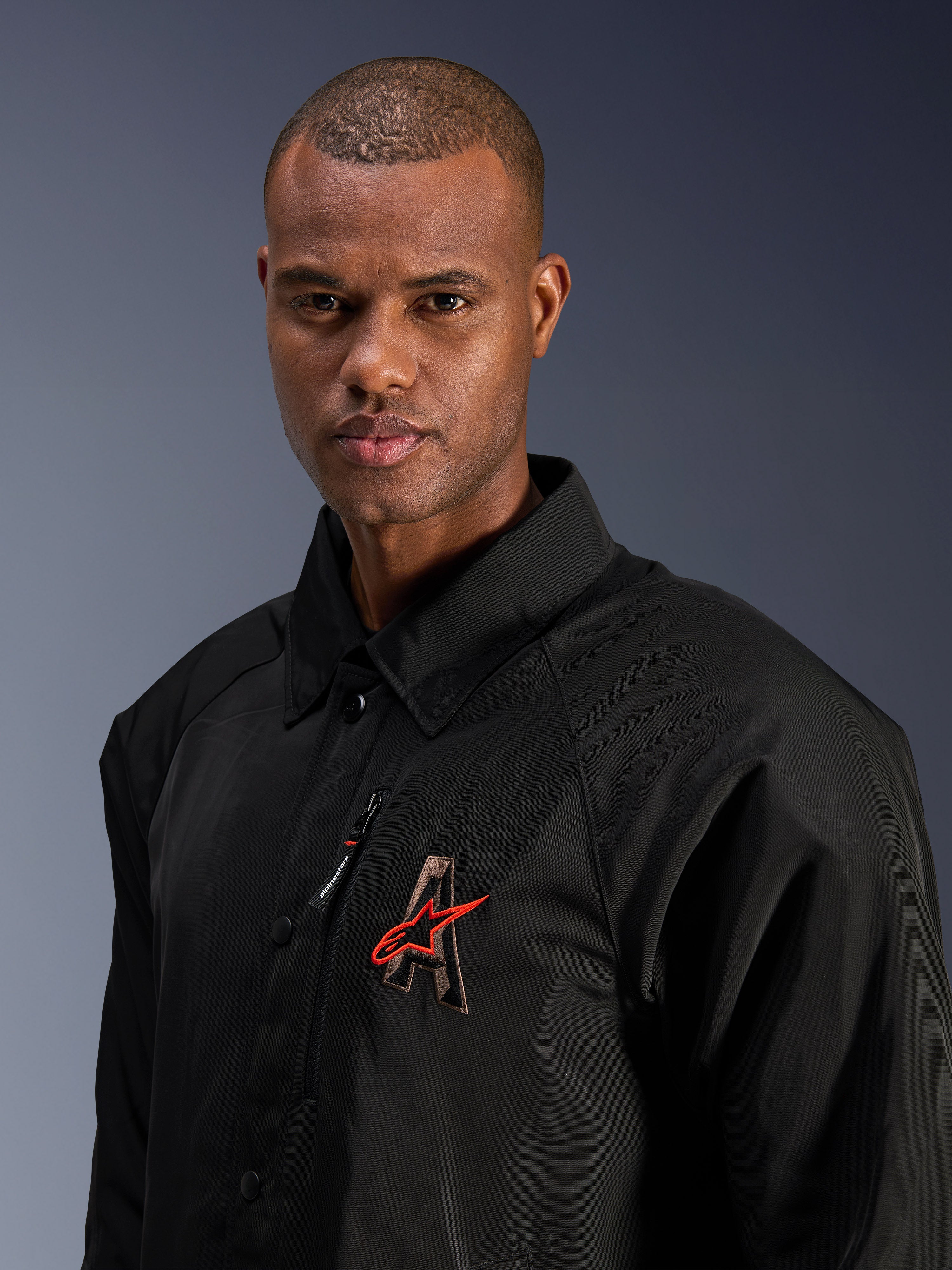 Veste Alpinestars Revved WR, noire, gros plan d'un homme portant la veste de moto en satin résistante à l'eau, avec le logo « A » brodé en 3D et l'étoile Alpinestars rouge, une poche de poitrine zippée et un design avec col pour le pilotage urbain contemporain.