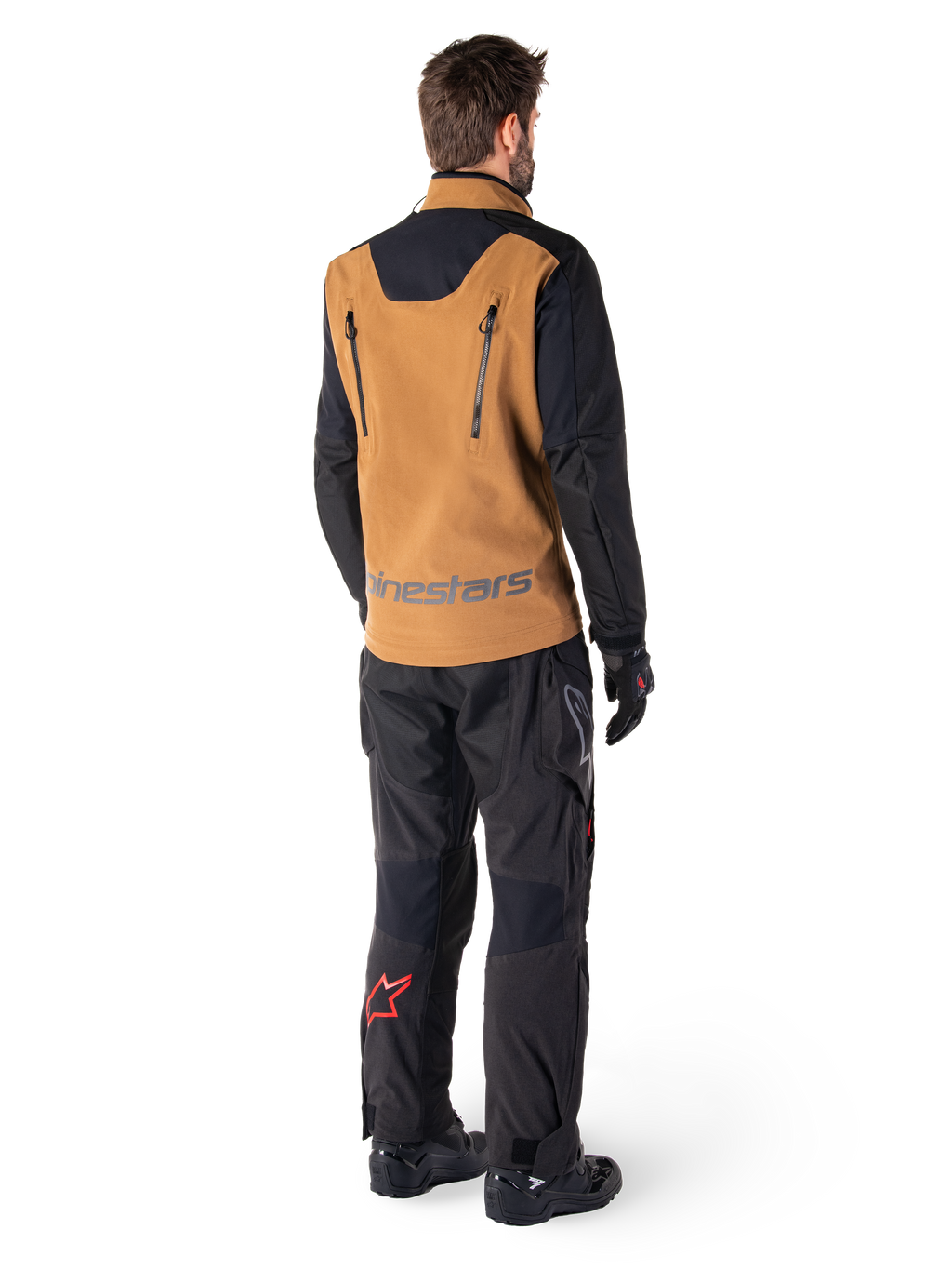 Veste Alpinestars Hyde XT Stretch Drystar®XF et pantalon Hyde XT Drystar®, vue arrière d'un pilote portant un équipement de touring aventure beige et noir, veste dotée de fermetures éclair verticales dans le dos et du marquage Alpinestars, pantalon noir avec logo étoile rouge sur le mollet, associé à des gants et des bottes noirs.