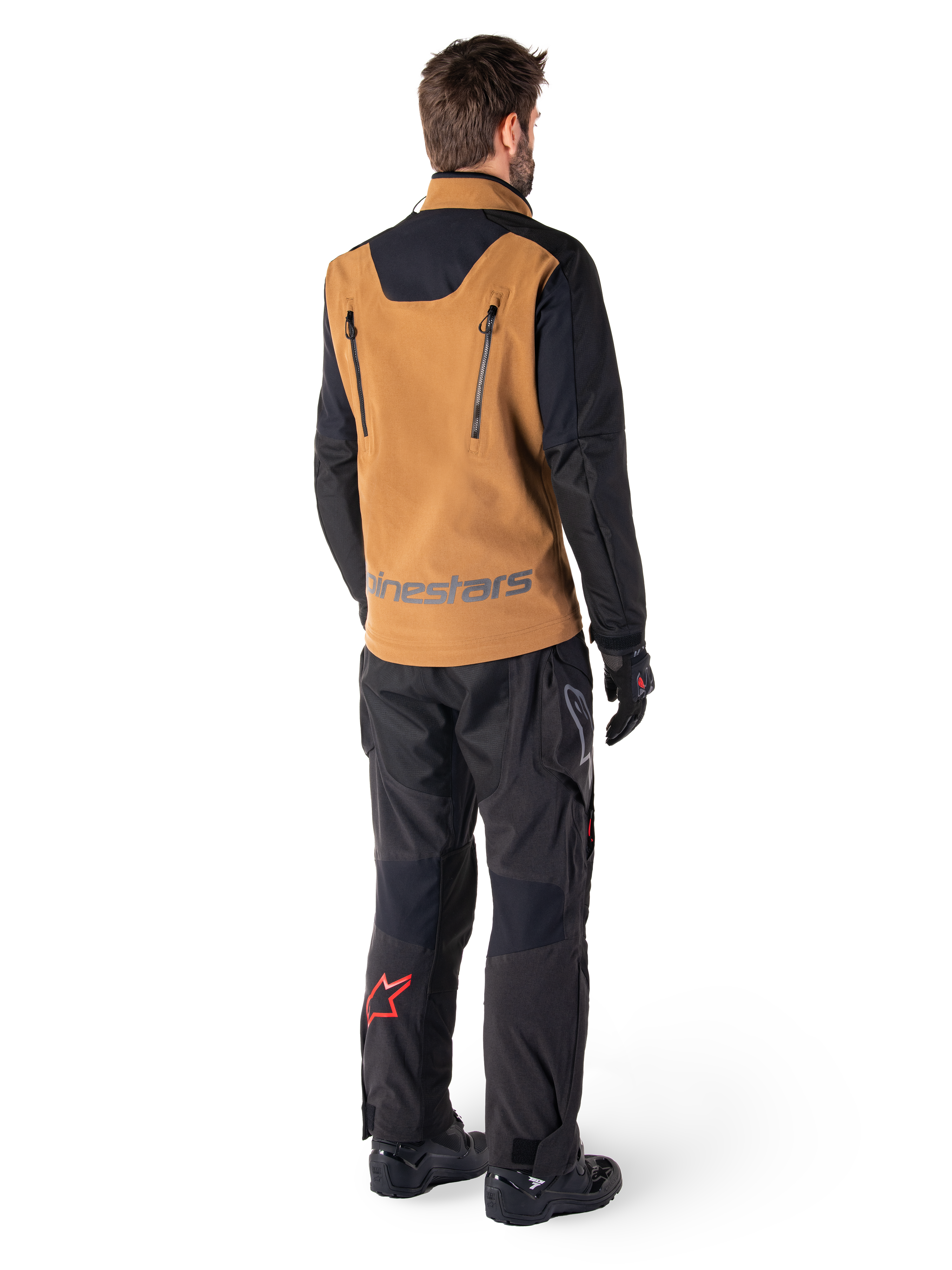 Veste Alpinestars Hyde XT Stretch Drystar®XF et pantalon Hyde XT Drystar®, vue arrière d'un pilote portant un équipement de touring aventure beige et noir, veste dotée de fermetures éclair verticales dans le dos et du marquage Alpinestars, pantalon noir avec logo étoile rouge sur le mollet, associé à des gants et des bottes noirs.