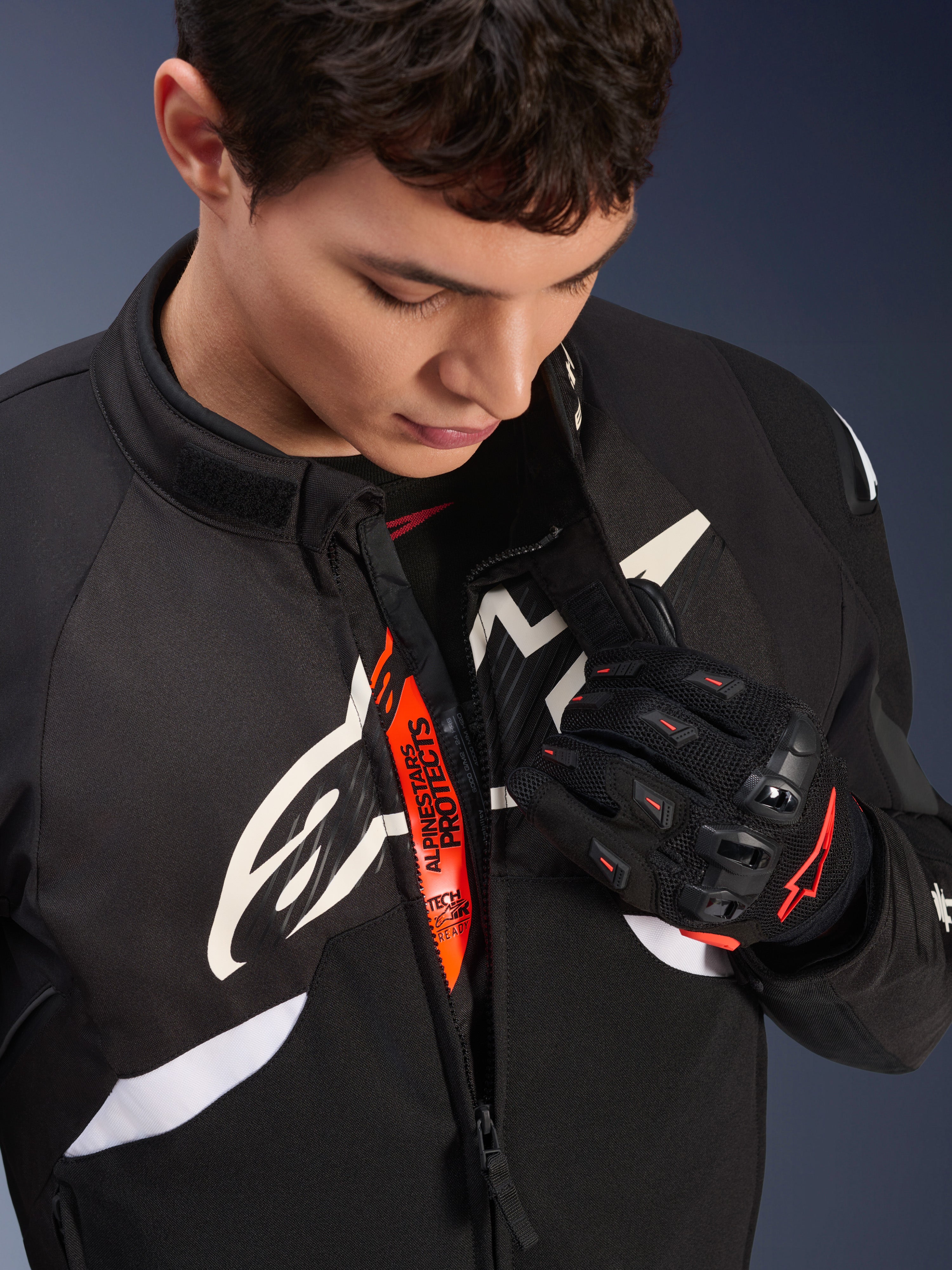 Alpinestars T-GP R V3 Drystar® Jacket, Black and White motorcycle sport textile jacket, partially unzipped revealing orange 'Alpinestars Protects' inner strap, worn with black and red mesh gloves, race-derived protective apparel.