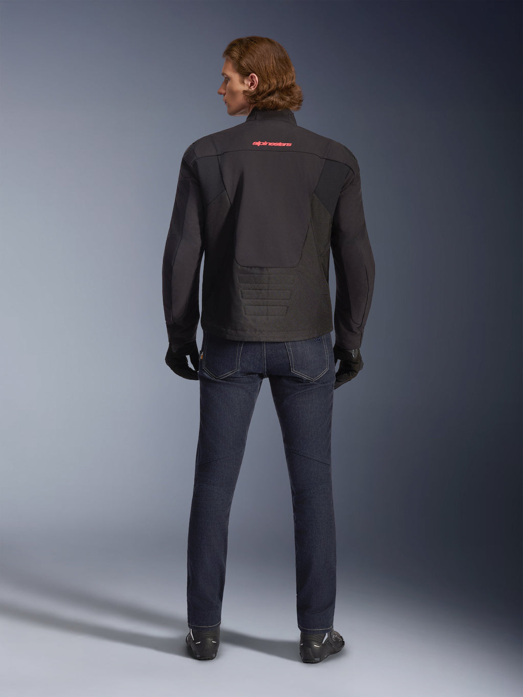 Veste imperméable Alpinestars SMX, noire, vue de dos portée par un mannequin, arborant un logo rouge en haut du dos, ourlet arrière allongé pour une meilleure couverture en conduite, assortie à un jean de moto bleu foncé et des gants noirs.