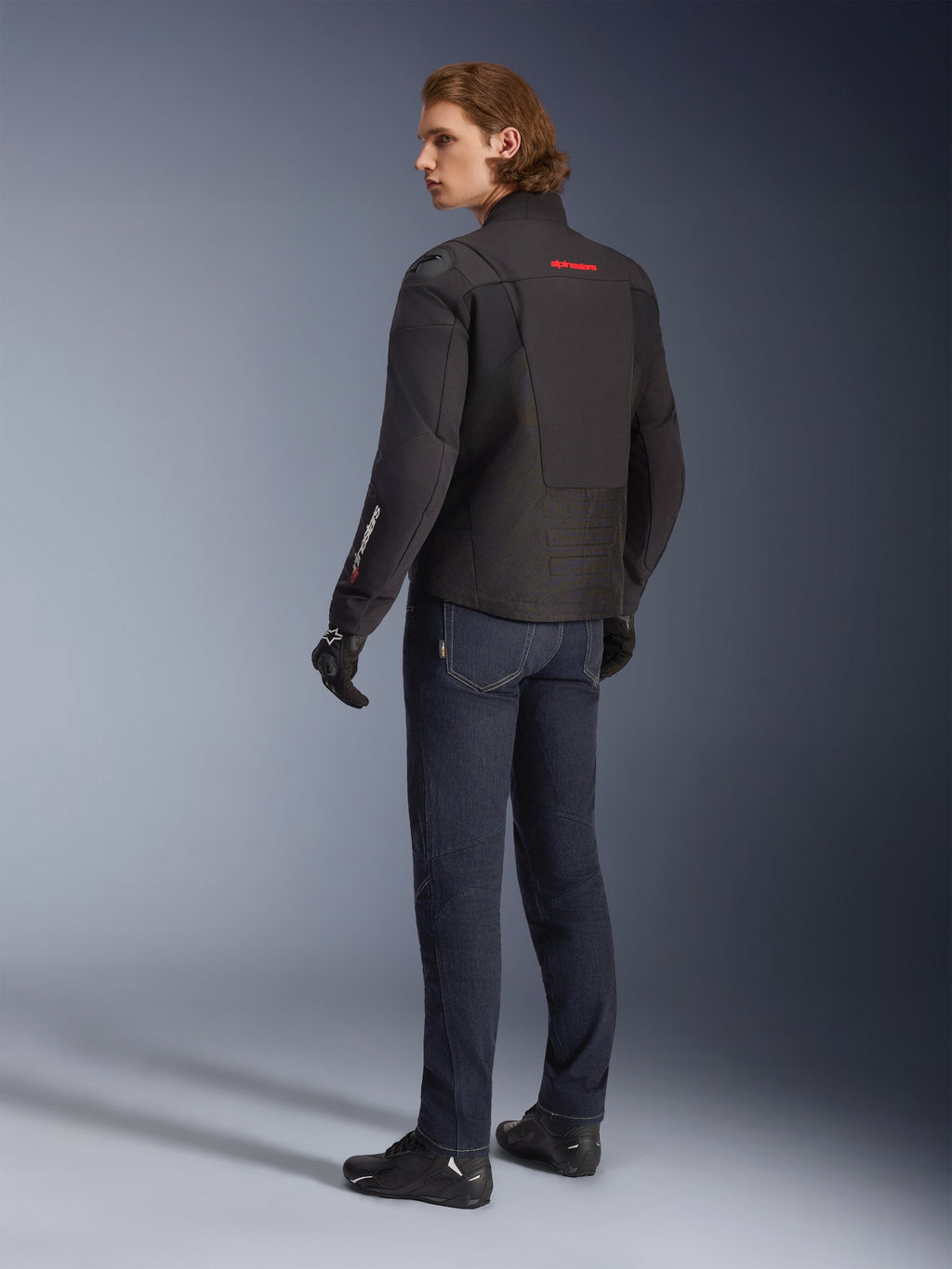 Veste imperméable Alpinestars SMX, veste de moto noire avec des touches de rouge et de blanc, motard vu de dos portant un jean de moto bleu foncé et des gants noirs, dotée de protections extérieures aux épaules et d'un pan arrière allongé pour la conduite sur route