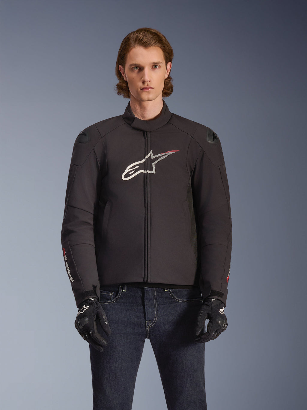 Alpinestars SMX Waterproof Jacket, veste de moto en textile noir avec logo étoile blanc et rouge sur la poitrine, portée par un mannequin avec des gants noirs et un jean bleu, dotée de sliders d'épaule externes et d'une coupe sportive.