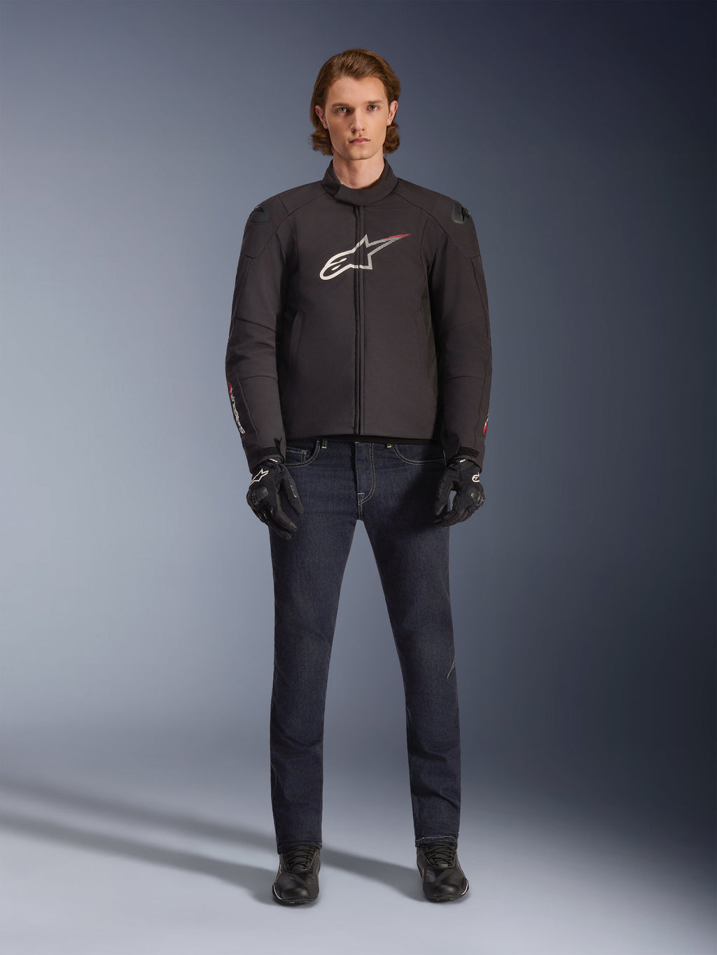 Modèle portant la veste imperméable Alpinestars SMX en noir avec logo blanc et rouge, associée à un pantalon de moto en denim foncé, des gants noirs et des bottes de moto, tenue de moto sportive pour toutes les conditions météorologiques.