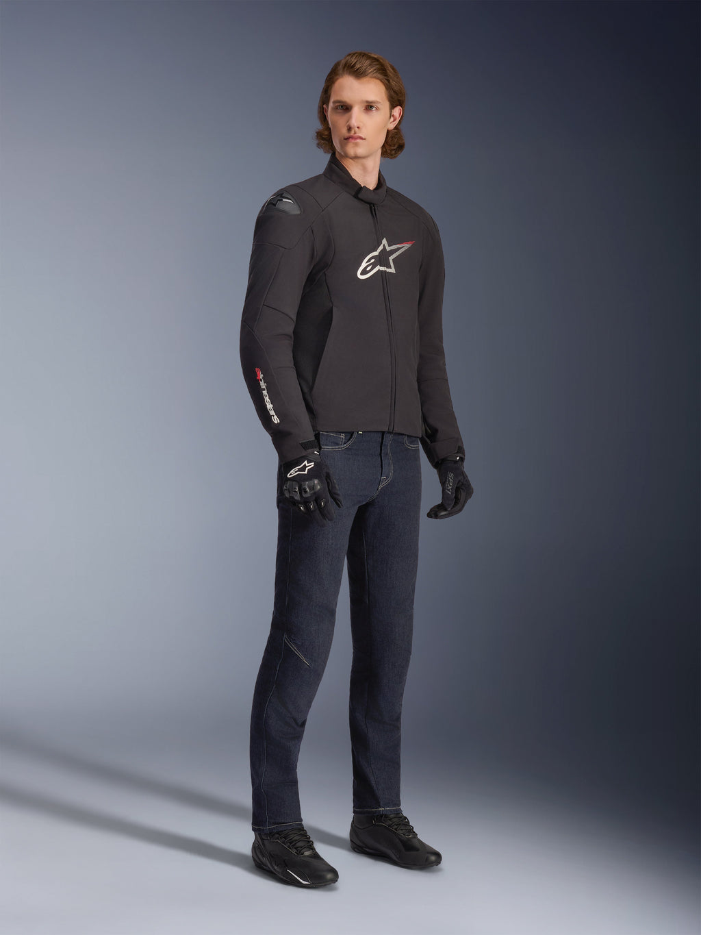 Modèle masculin portant la veste imperméable Alpinestars SMX, noire avec des accents de logo blancs et rouges, associée à un pantalon en jean bleu foncé et des gants de moto noirs, veste de moto en textile élégante à coupe sportive.