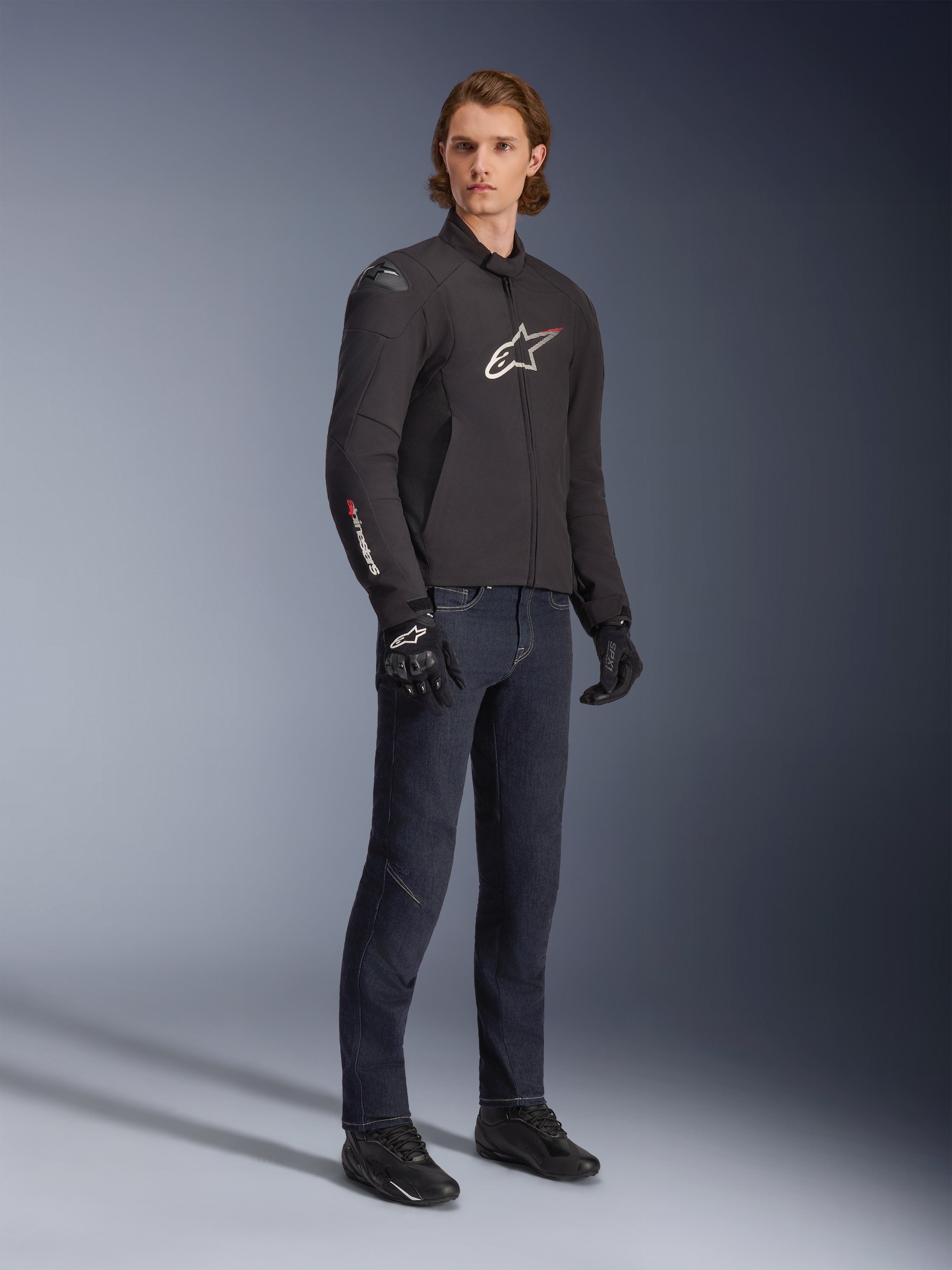 Modèle masculin portant la veste imperméable Alpinestars SMX, noire avec des accents de logo blancs et rouges, associée à un pantalon en jean bleu foncé et des gants de moto noirs, veste de moto en textile élégante à coupe sportive.