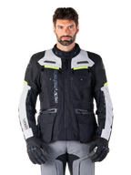 Homme portant une veste Alpinestars Bogotá Pro Drystar®, noir, gris glacé et jaune fluo, veste de moto touring aventure toutes saisons, conception imperméable avec plusieurs poches et marquage vertical, associée à des gants noirs et un pantalon de moto gris