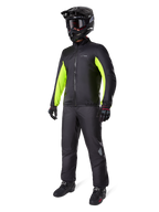 Motard portant la doublure de la veste Alpinestars Bogotá Pro Drystar®, noire avec des empiècements jaune fluo sur les manches, accompagnée d'un pantalon noir assorti, d'un casque de motocross noir et de gants, équipement d'aventure imperméable toutes saisons