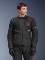 Veste Alpinestars Andes Air Drystar®, veste de moto touring noire, logo étoile rouge sur la poitrine, avec panneaux de ventilation en mesh et détails réfléchissants, portée par un mannequin homme avec des gants de moto noirs, équipement d'aventure deux saisons