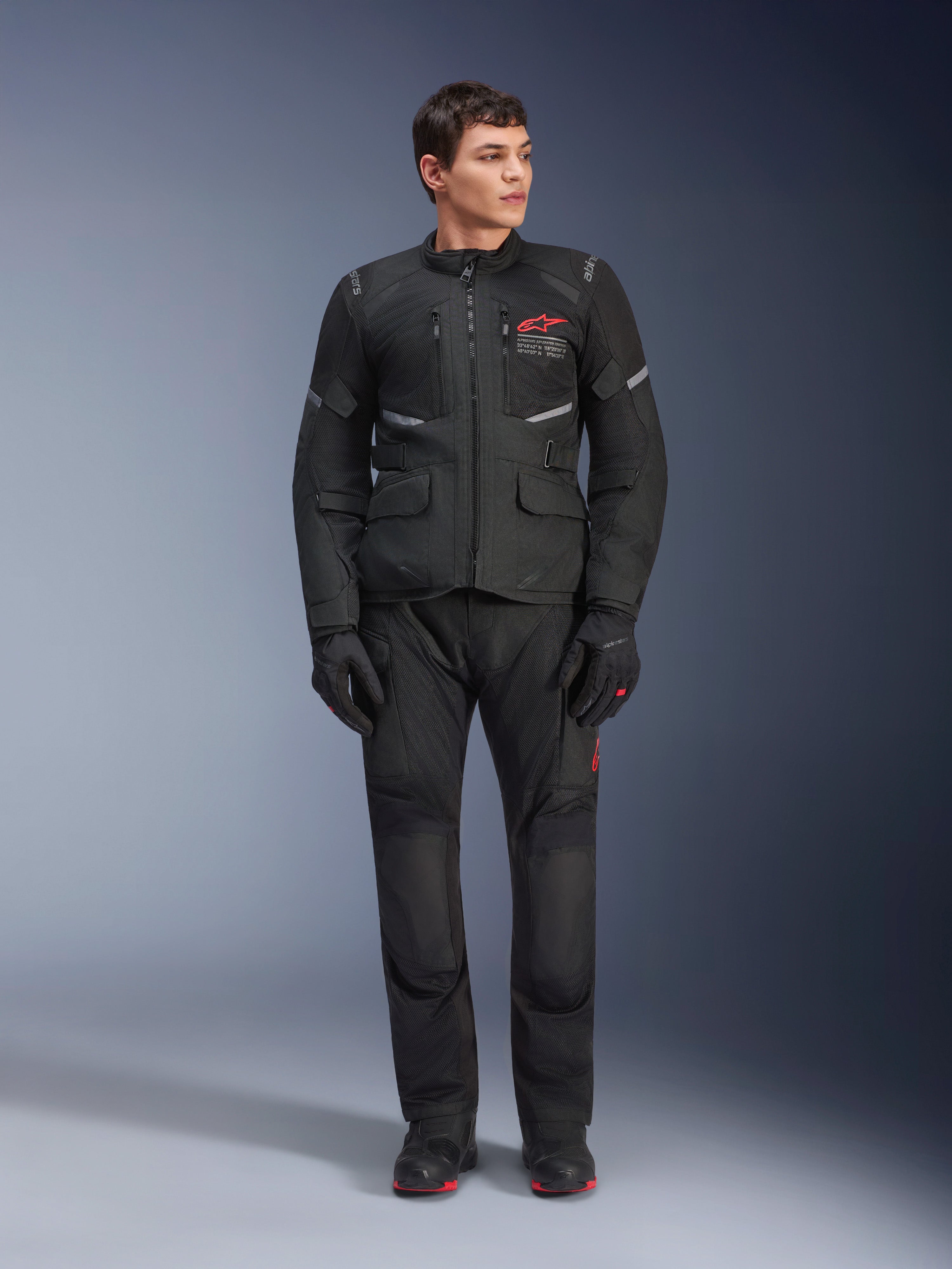 Modèle masculin portant la veste Alpinestars Andes Air Drystar® noire, équipement de moto aventure touring avec pantalon et gants assortis, avec logo rouge sur la poitrine, ventilation en maille et poches fonctionnelles.