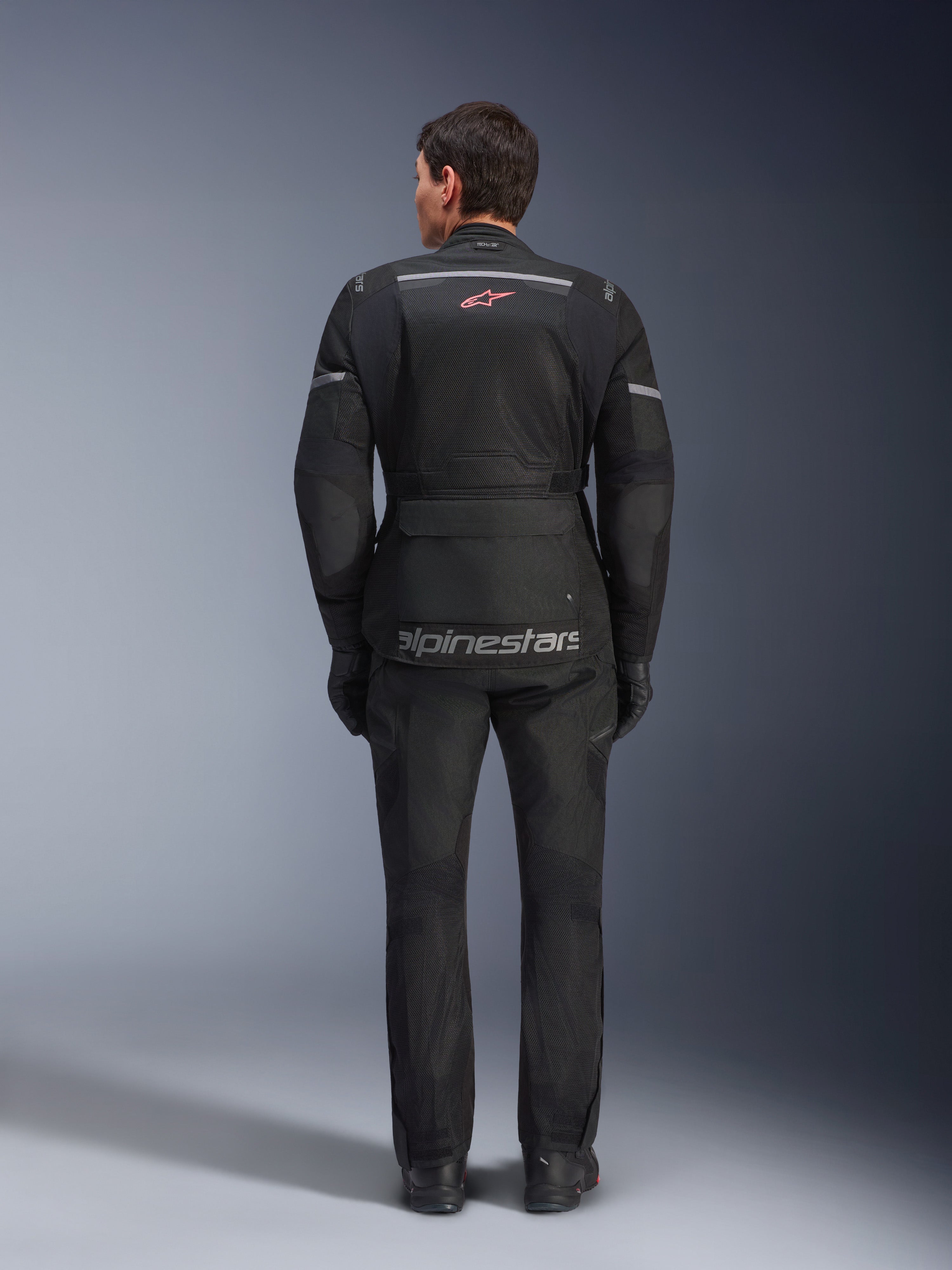 Vue arrière d'un pilote portant la veste Alpinestars Andes Air Drystar® noire, dotée de larges panneaux de ventilation en mesh au dos, d'un logo Alpinestars rouge et de détails réfléchissants, associée à un pantalon et des gants de moto assortis pour le touring aventure.