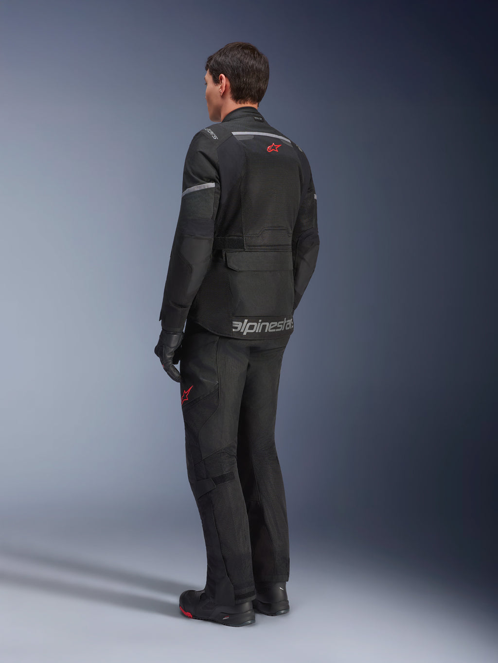 Veste Alpinestars Andes Air Drystar®, Noir, vue arrière sur un motard, dotée de larges panneaux en mesh pour la ventilation, portée avec un pantalon de moto d'aventure et des bottes assortis, mettant en avant le design de touring orienté route et l'image de marque.