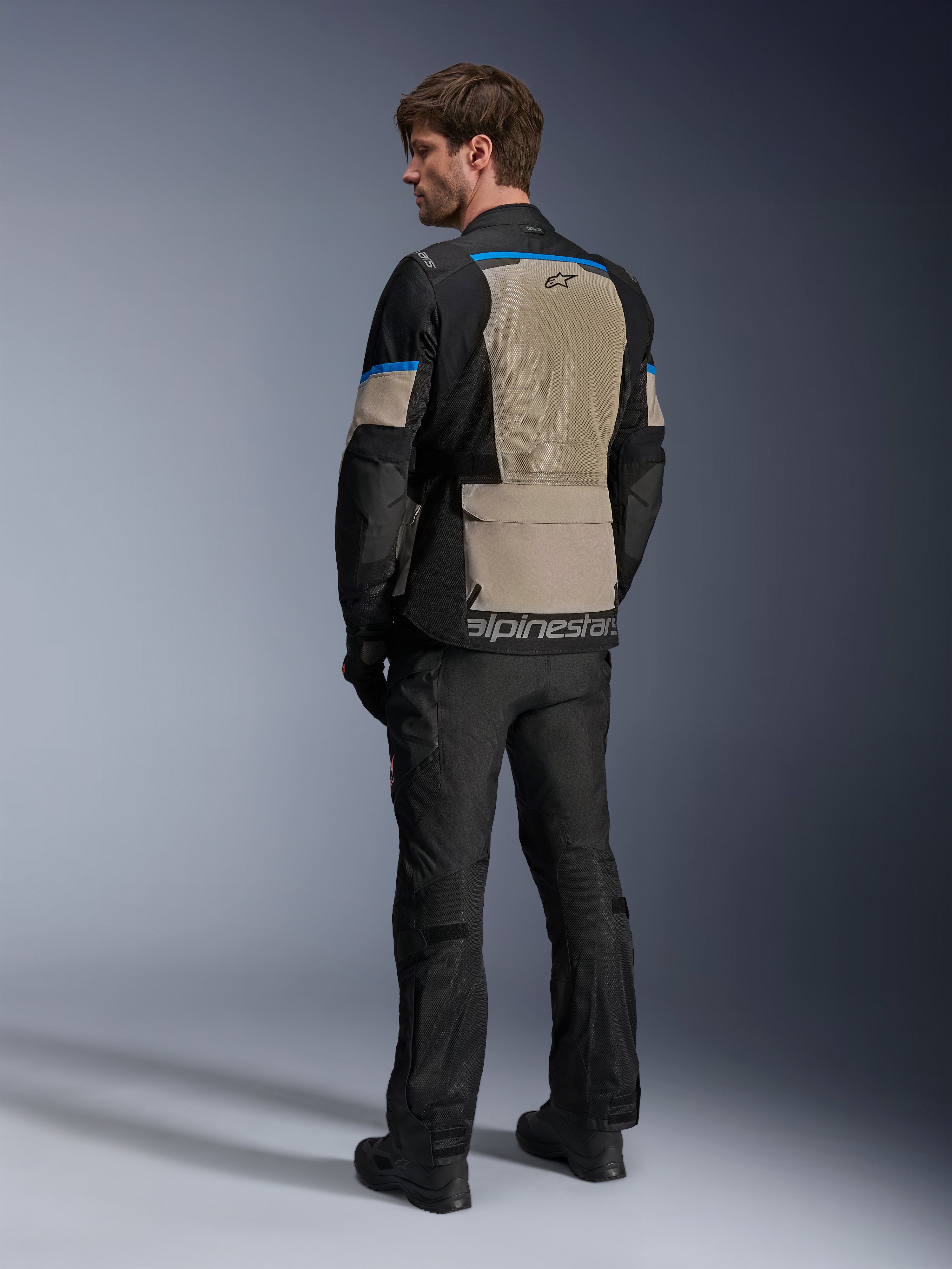 Veste Alpinestars Andes Air Drystar®, noir, gris laurel et bleu vif, vue arrière montrant un large panneau de ventilation en maille dans le dos, logo sur l'ourlet inférieur, portée avec un pantalon et des bottes de moto noirs.