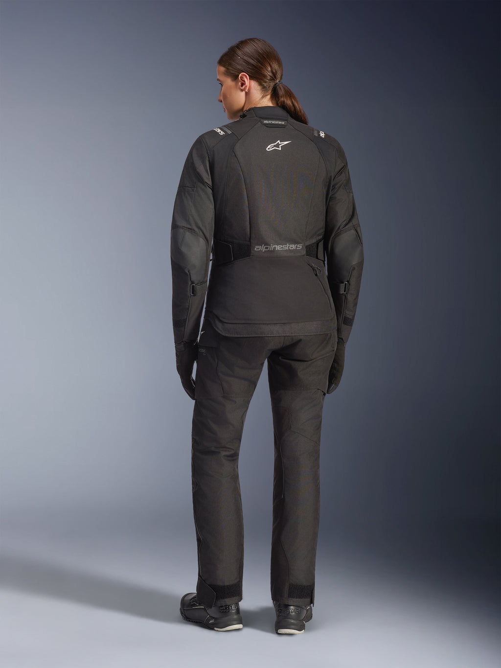 Motarde vue de dos portant la veste Alpinestars Stella Andes V4 Drystar® noire, associée à un pantalon de moto assorti, logo Astar blanc dans le dos, ourlet arrière allongé, tenue d'aventure-touring complète incluant gants et bottes noirs