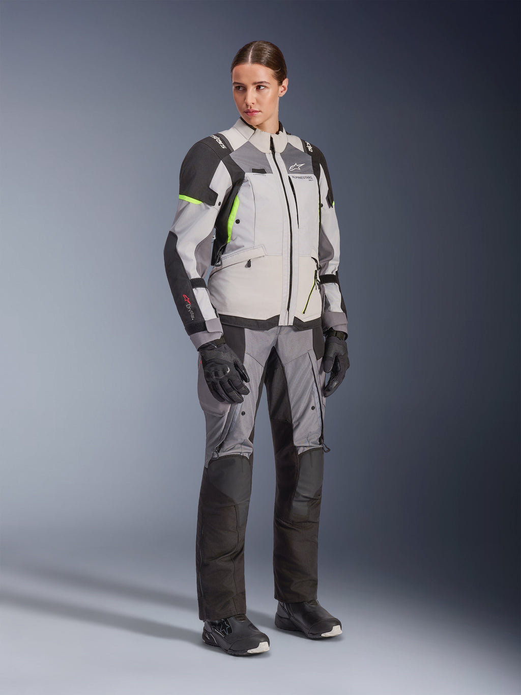 Femme portant la veste Alpinestars Stella Andes V4 Drystar® et le pantalon assorti, Gris Glace et Gris Foncé avec des accents Jaune Fluo, ensemble complet d'équipement de touring aventure avec gants et bottes noirs, photo en pied en studio.