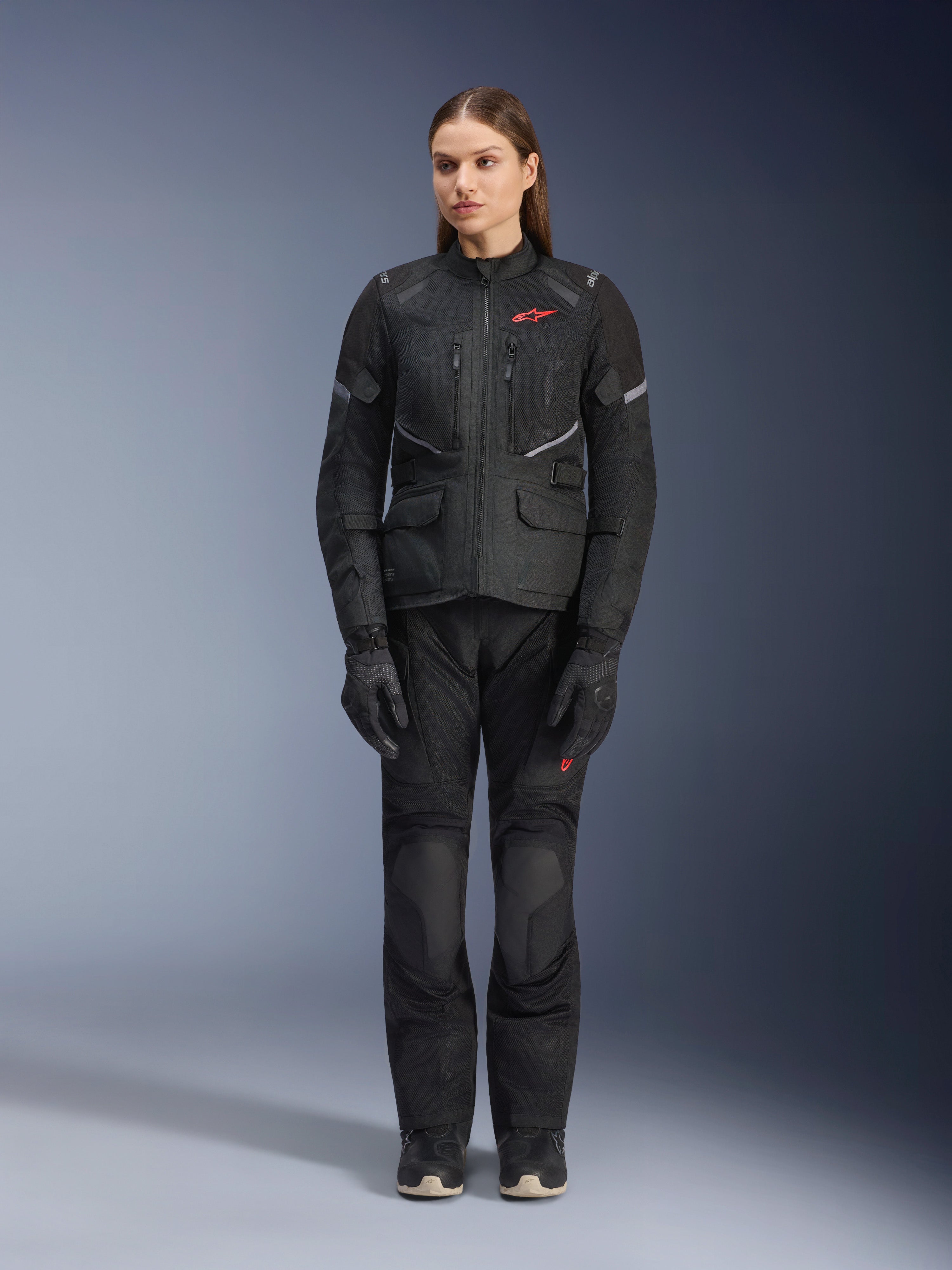Modèle féminin portant la veste Alpinestars Stella Andes Air Drystar® noire, assortie d'un pantalon de touring et de gants noirs, avec de larges panneaux de ventilation en maille, des touches de logo rouge et des détails réfléchissants, photo de studio professionnelle pour équipement de moto pour femme.
