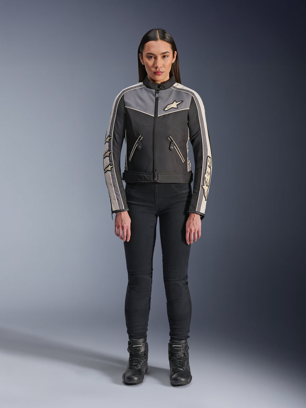 Femme portant la veste Alpinestars Stella T-Dyno WR, noir, gris foncé et écru, vue de face, veste de moto en textile au design d'inspiration vintage, avec logos sur la poitrine et les manches, portée avec un pantalon noir et des bottes