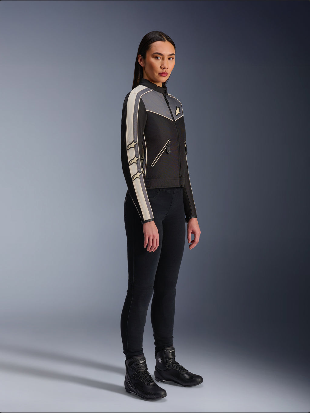 Veste Alpinestars Stella T-Dyno WR, veste de moto en textile pour femme, noir, gris foncé et écru, présentant un design d'inspiration vintage avec des bandes de manches crème et des logos étoiles, portée avec un pantalon noir et des bottes de moto, équipement de conduite urbaine d'inspiration héritage.