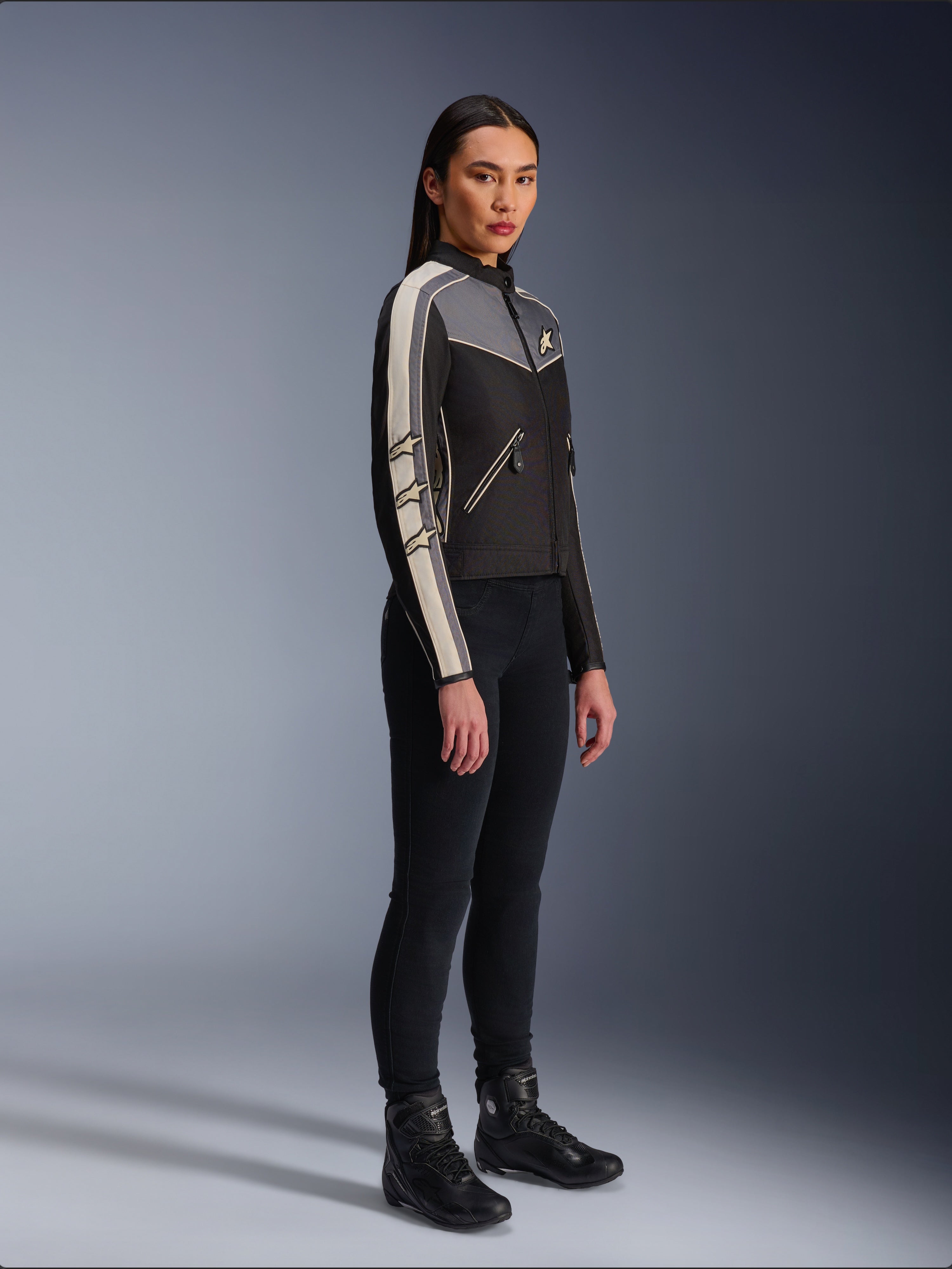 Veste Alpinestars Stella T-Dyno WR, veste de moto en textile pour femme, noir, gris foncé et écru, présentant un design d'inspiration vintage avec des bandes de manches crème et des logos étoiles, portée avec un pantalon noir et des bottes de moto, équipement de conduite urbaine d'inspiration héritage.