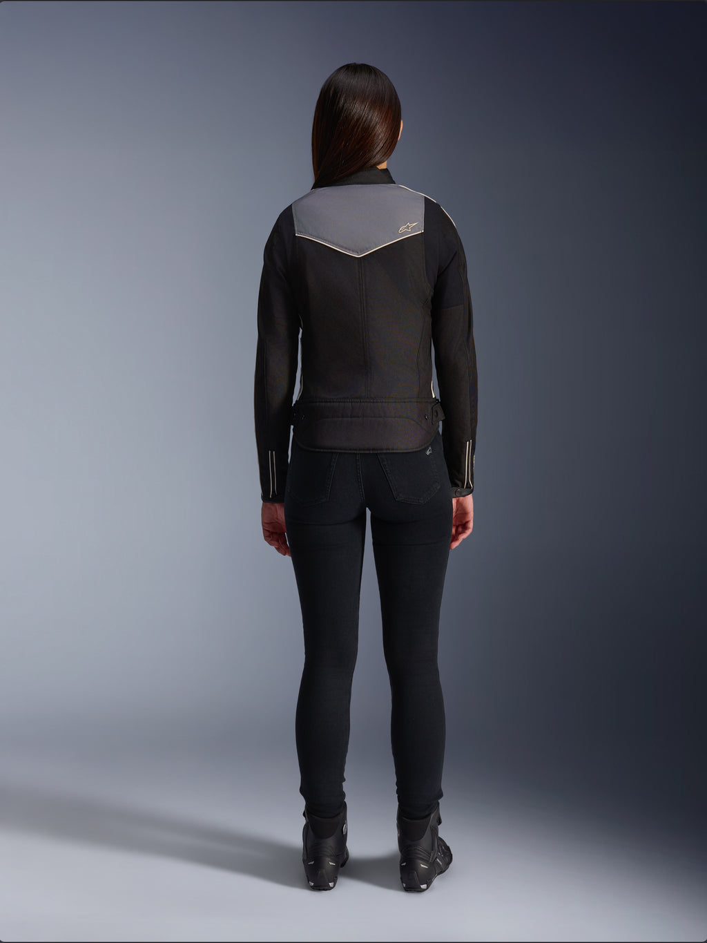Vue arrière d'une motarde portant la veste Alpinestars Stella T-Dyno WR en Noir, Gris Foncé et Écru, accompagnée d'un pantalon slim noir et de bottes de moto noires, mettant en valeur le panneau supérieur du dos gris avec le logo Alpinestars et les détails de finition écru.